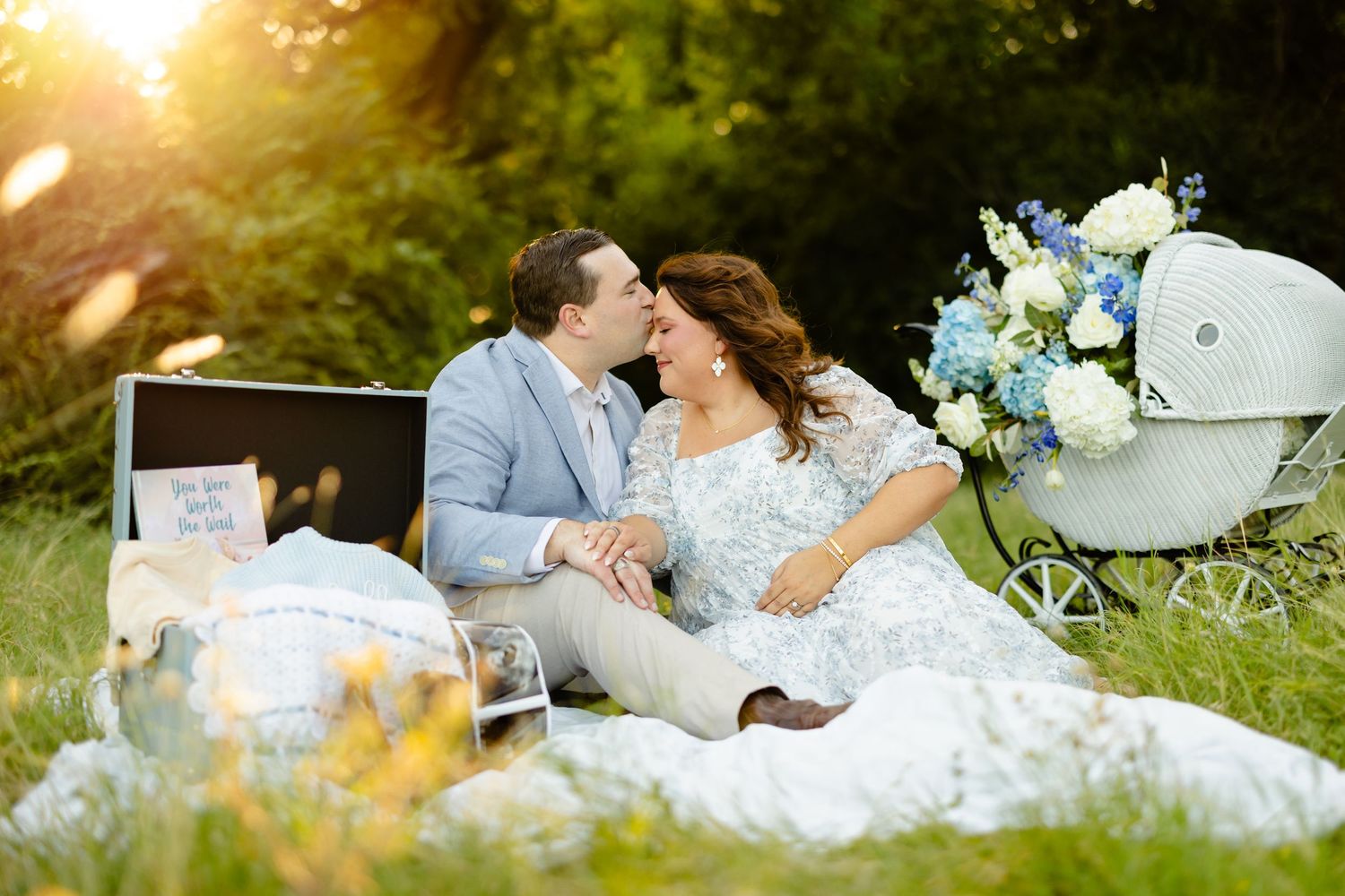 Couple sharing a romantic moment at a maternity photoshoot outdoors, with a vintage pram and blue floral bouquet.