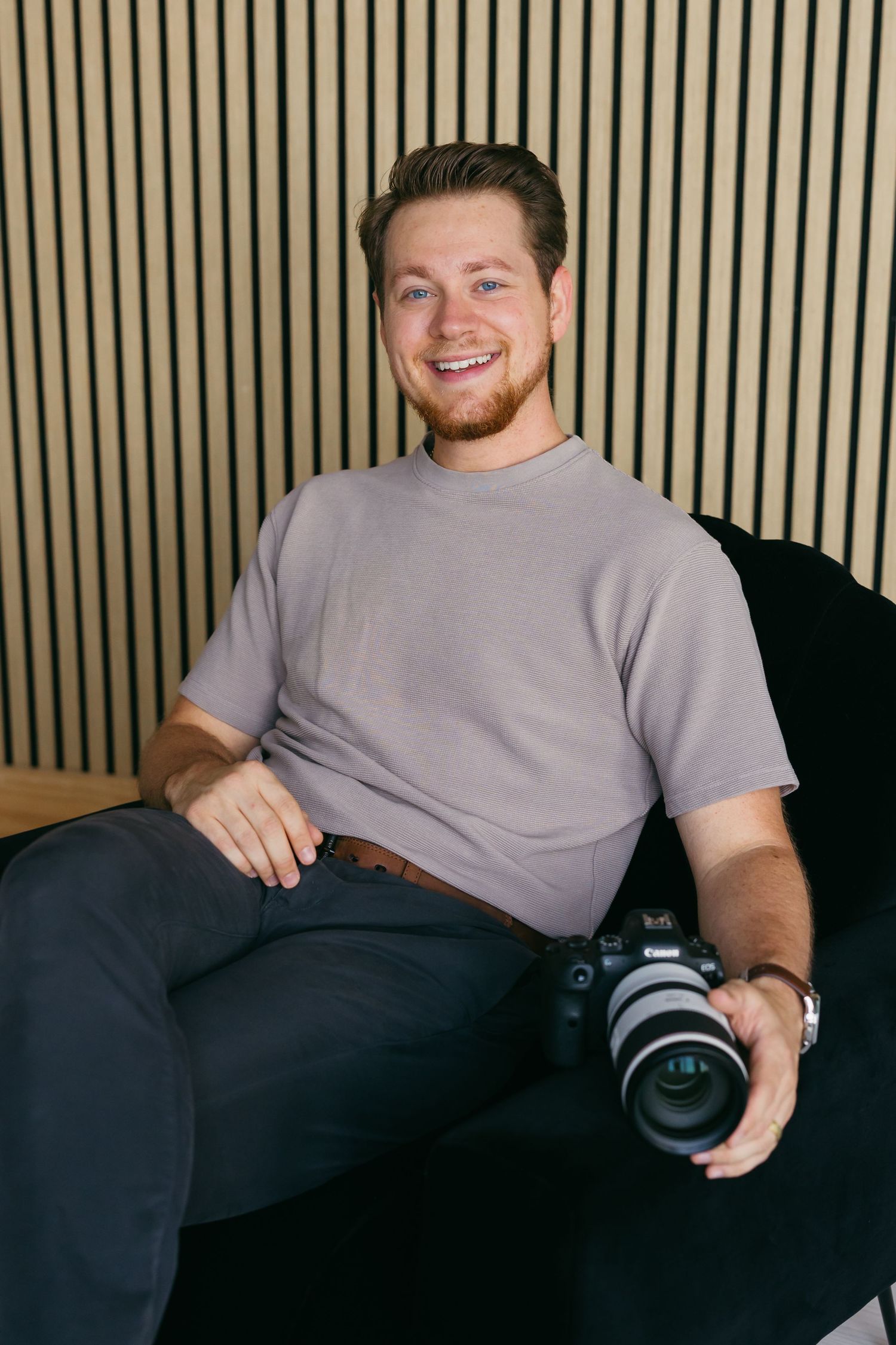 Person holding camera while sitting on dark couch against striped wall background.