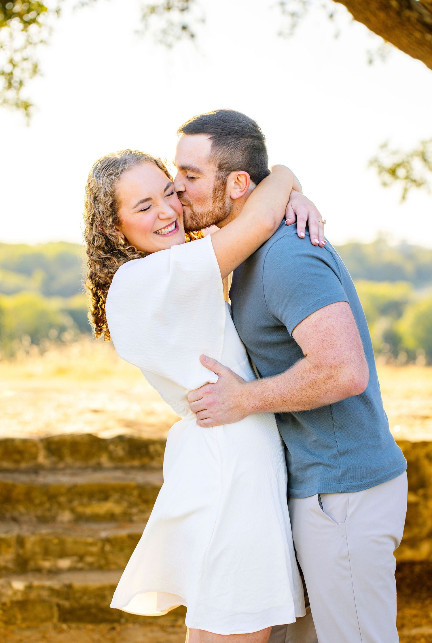 A romantic embrace between a newly engaged couple in white and blue casual wear during golden hour outdoors.