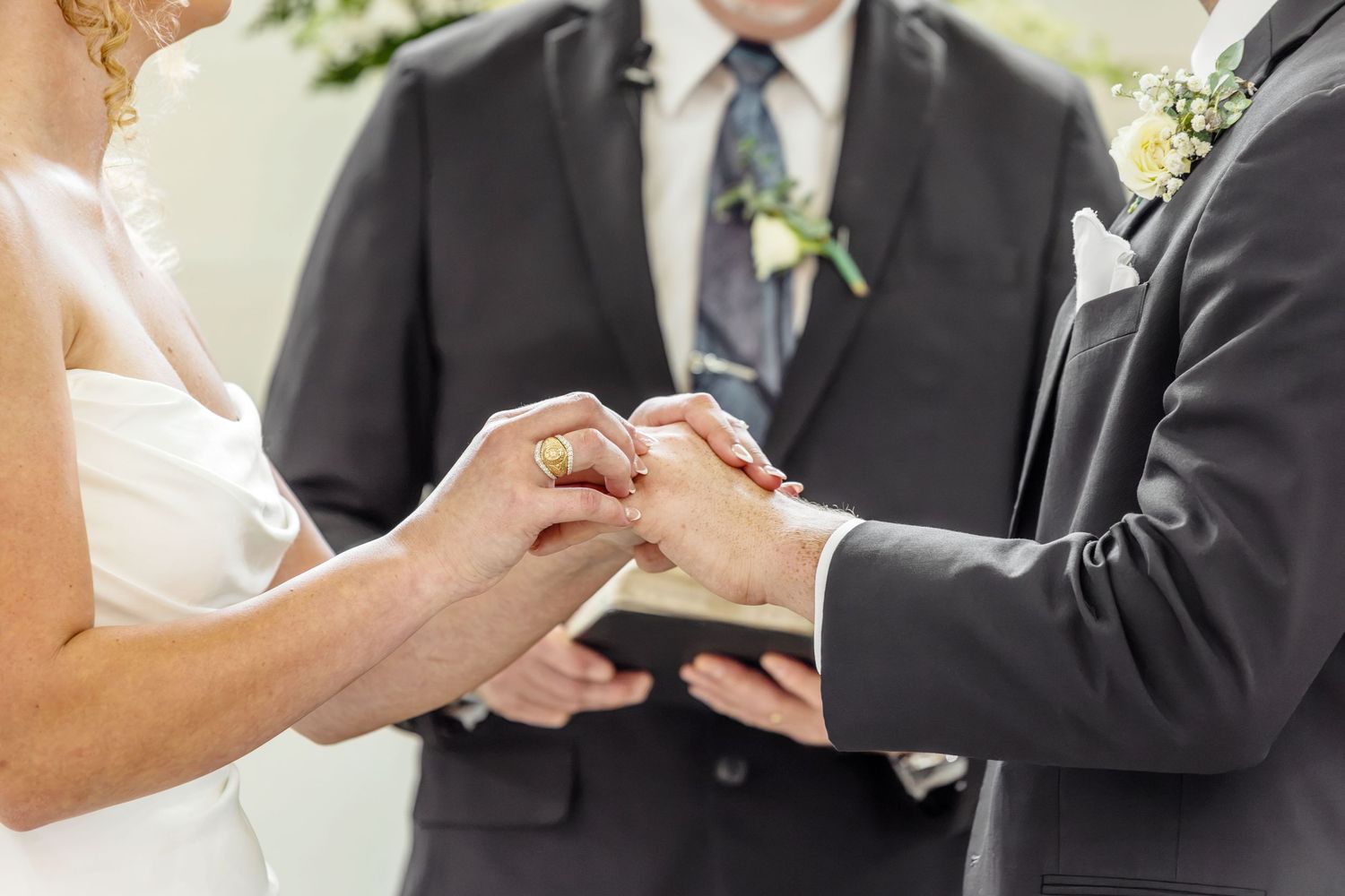 Bride and groom exchanging rings during wedding ceremony, officiant holding bible in background.