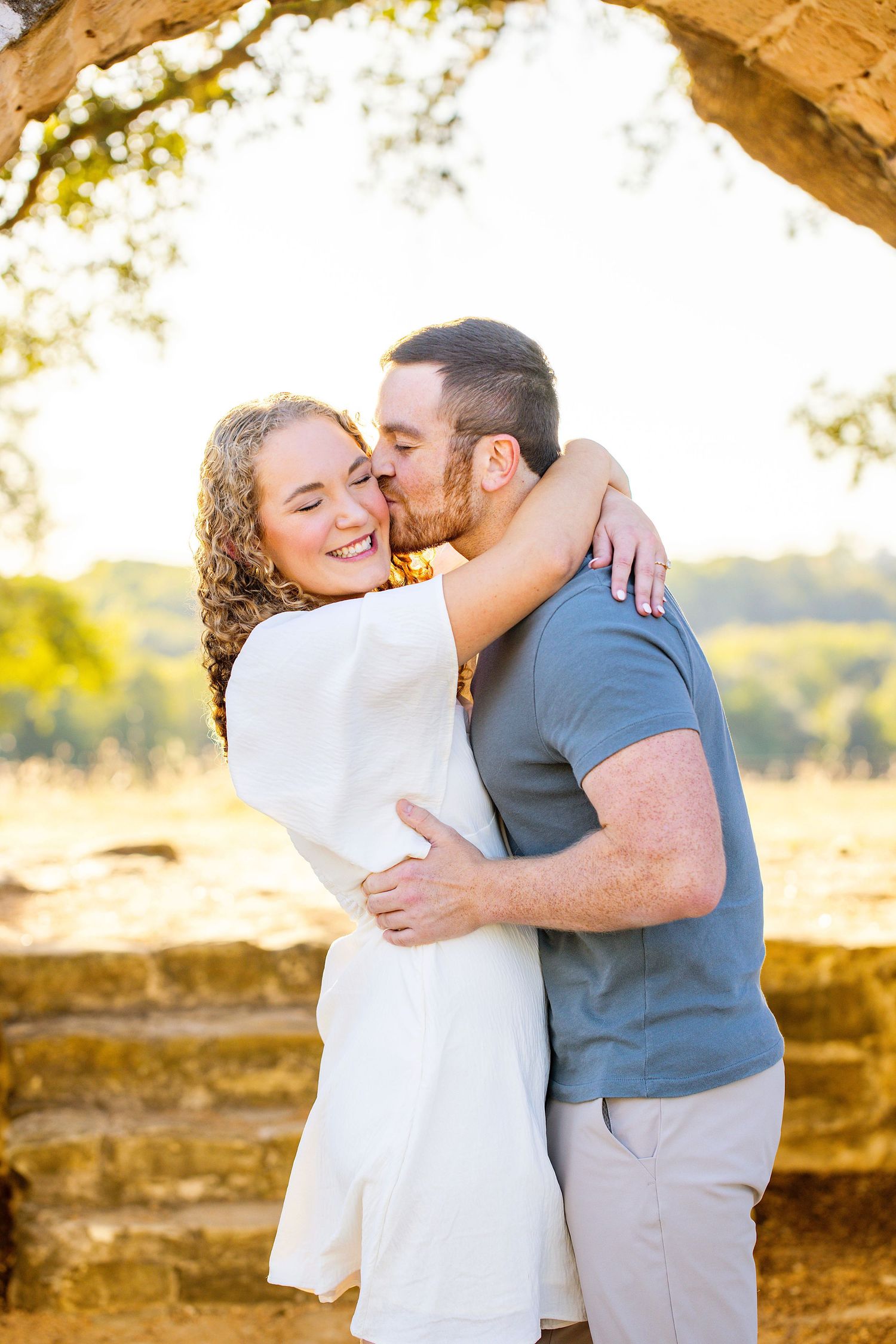 A couple embraces outdoors, woman in white dress laughing as man kisses her cheek, golden hour light.