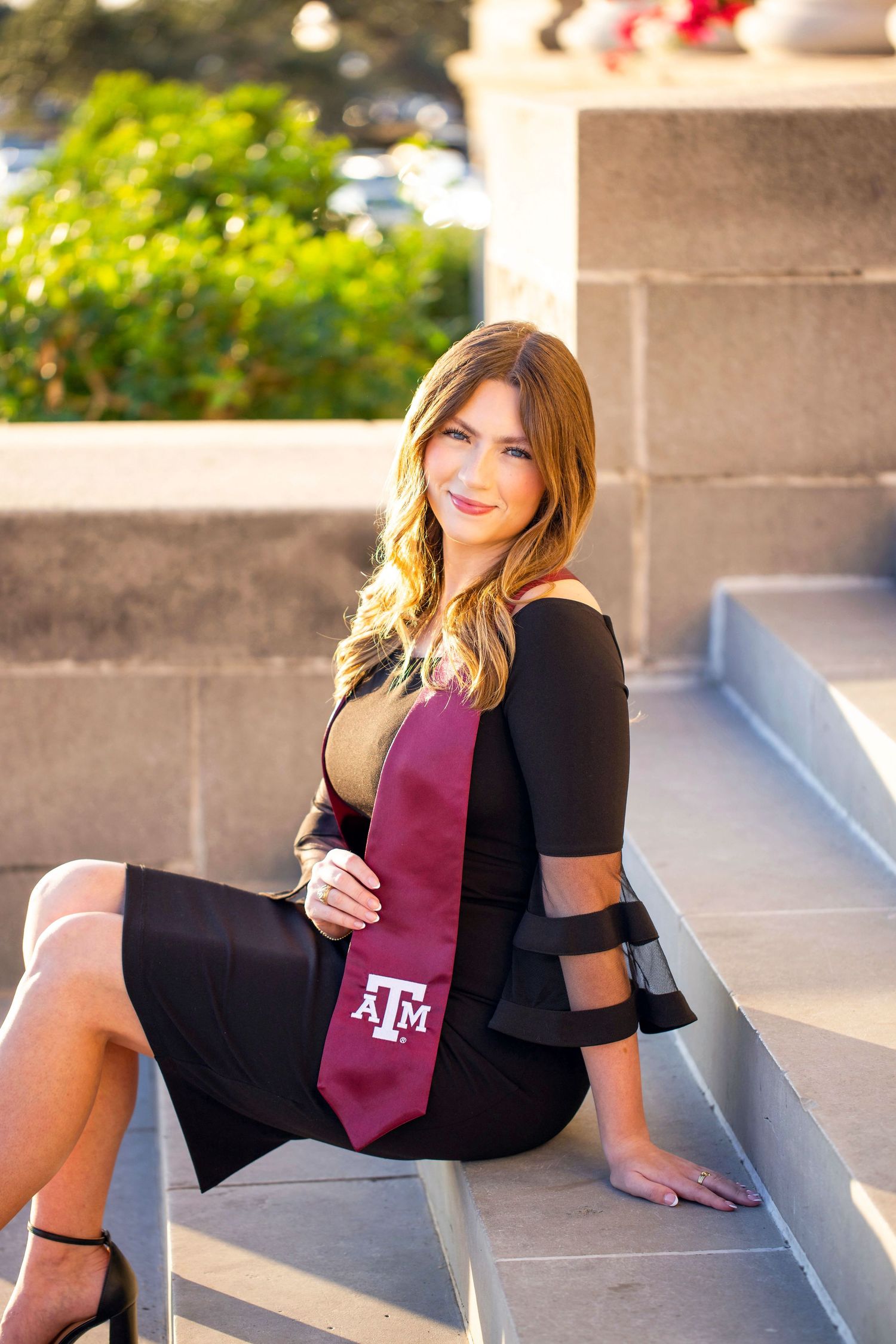 A young woman with long blonde hair wearing a black outfit and pink tie sits on concrete steps.