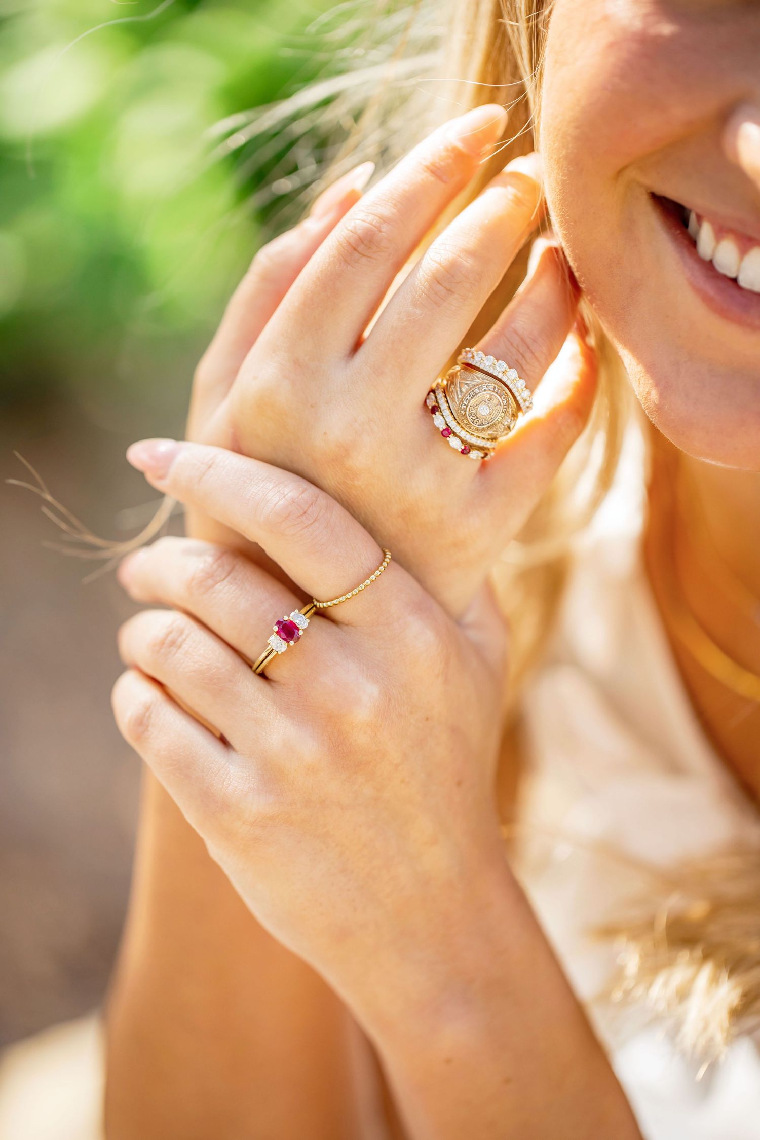 Woman smiling wearing gold rings with ruby gemstones and diamond accents on her fingers outdoors.
