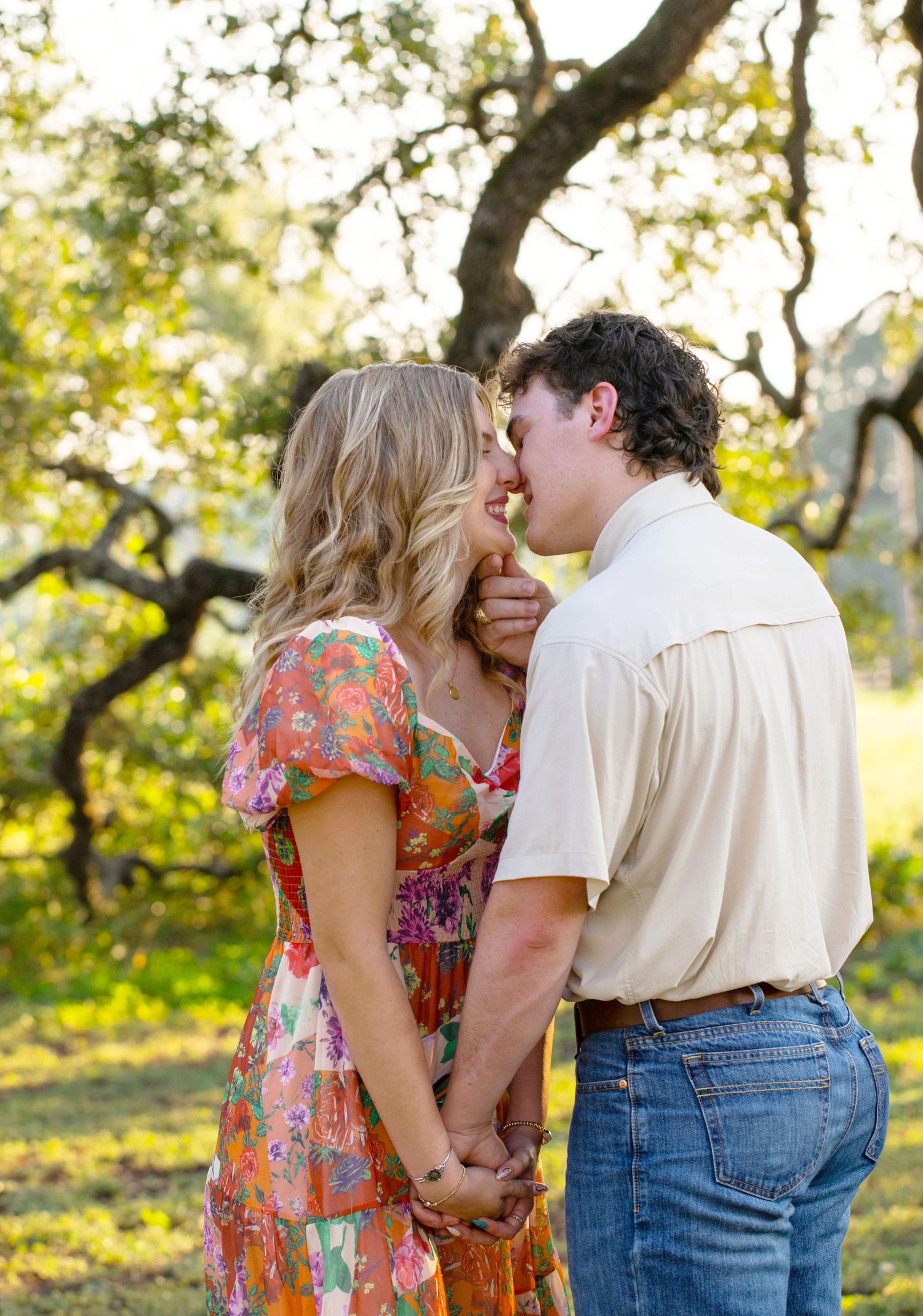 A couple shares a romantic moment in a floral dress and casual attire while standing in a sunlit orchard.