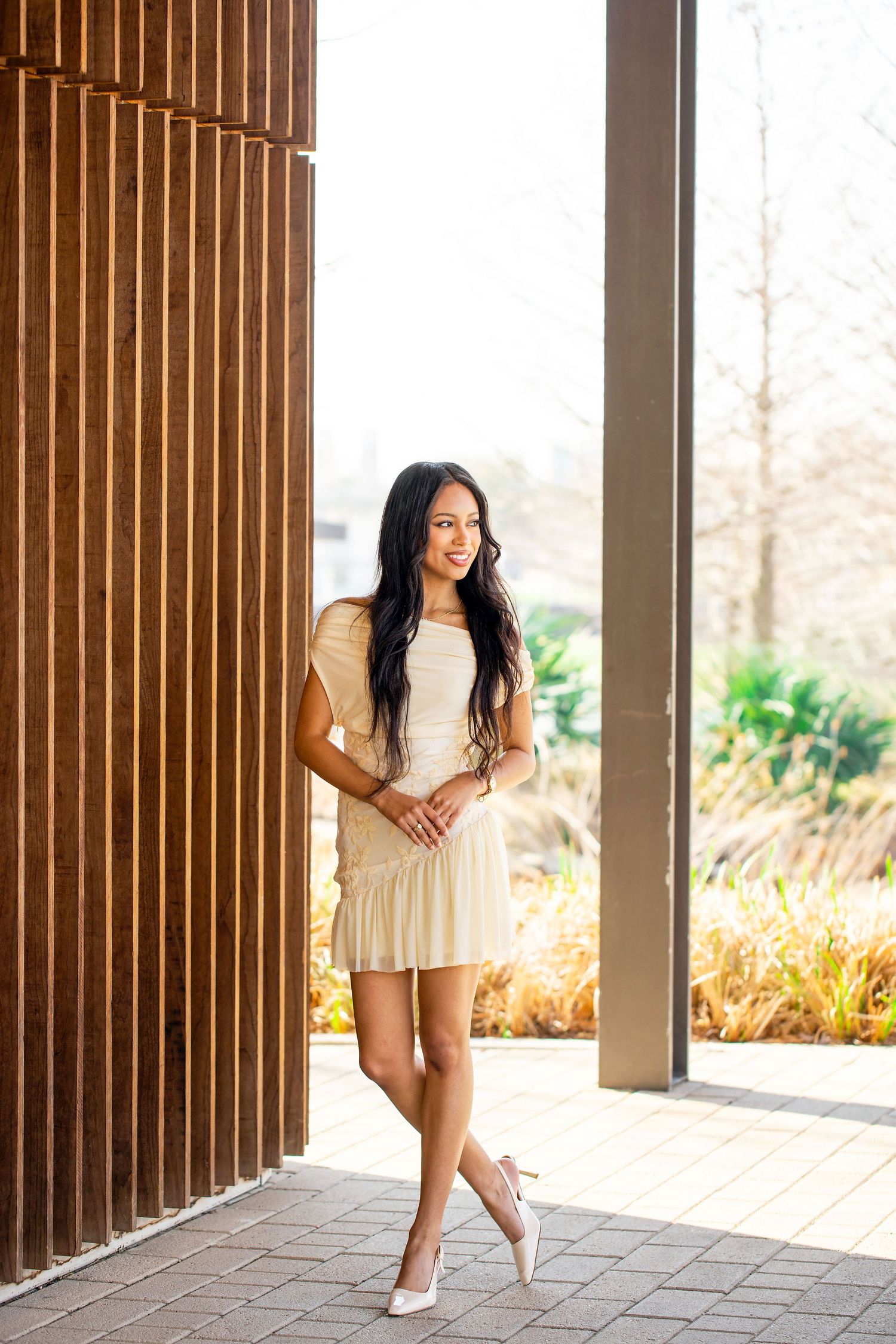 A woman in a beige dress poses confidently in front of wooden slats and natural lighting.