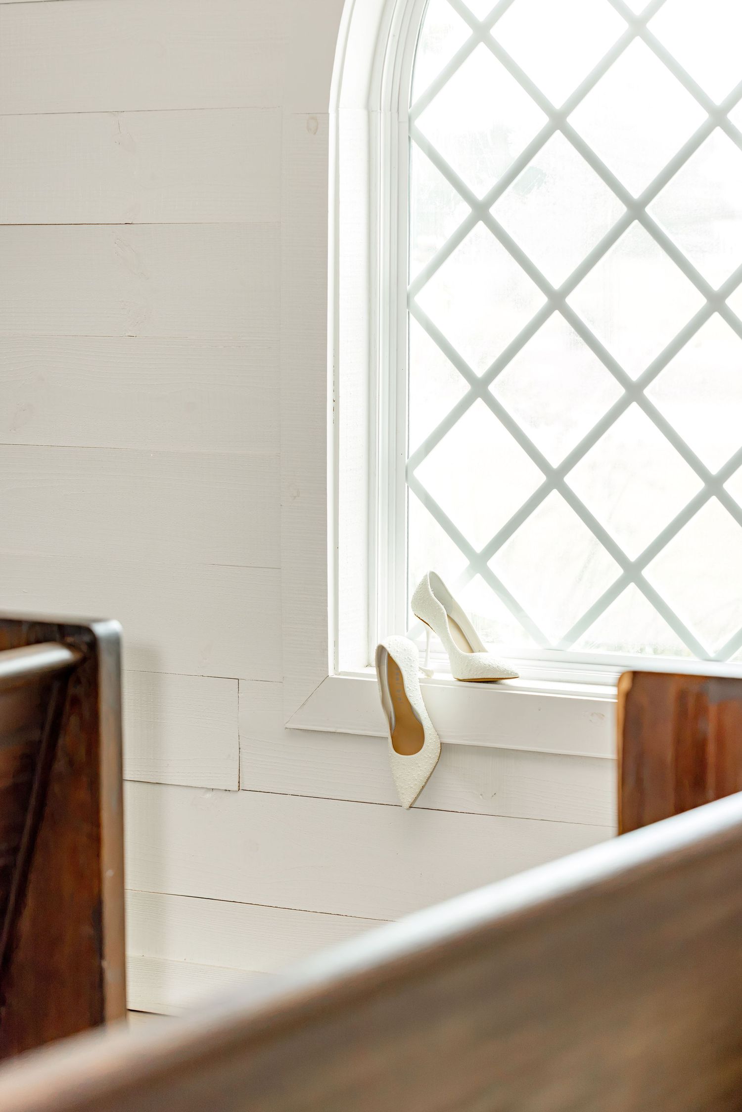 White bridal heels placed on a church windowsill with diamond-pane glass, beside wooden pews in a bright chapel.