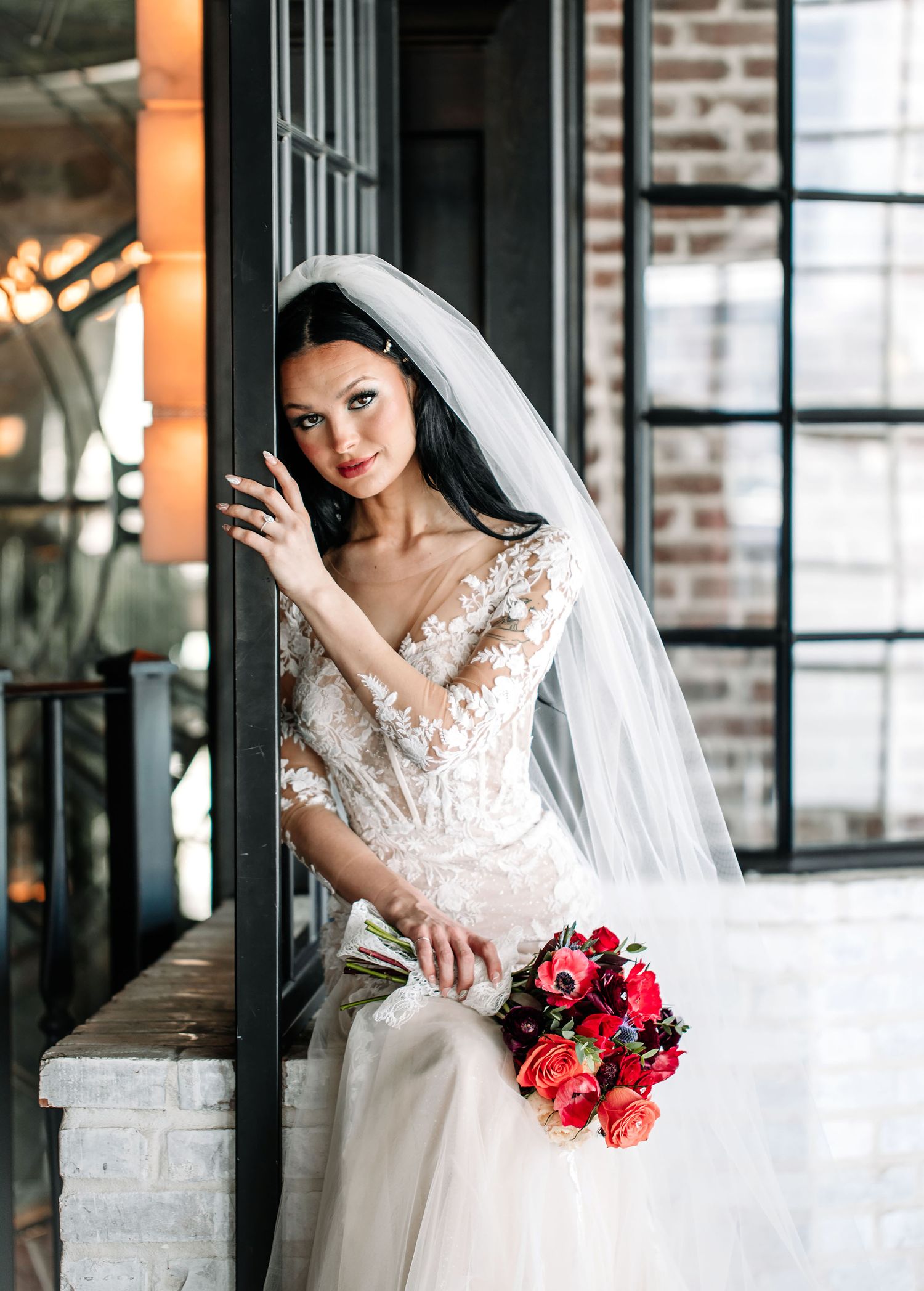 Beautiful bride in lace wedding dress with long veil poses by industrial window holding vibrant red and pink bouquet.