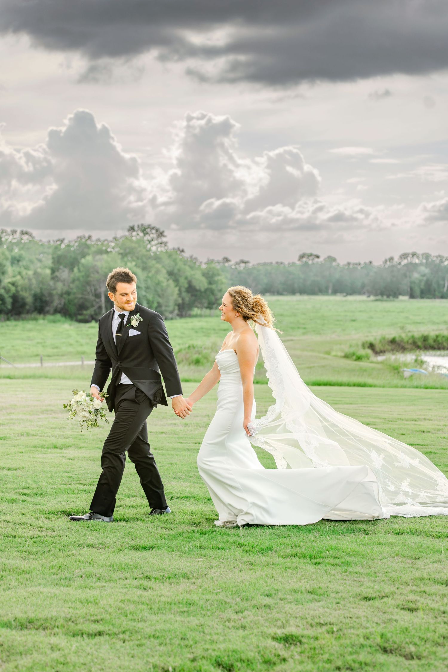 Bride and groom walk hand-in-hand across lush green field, her veil flowing in wind beneath dramatic cloudy sky.