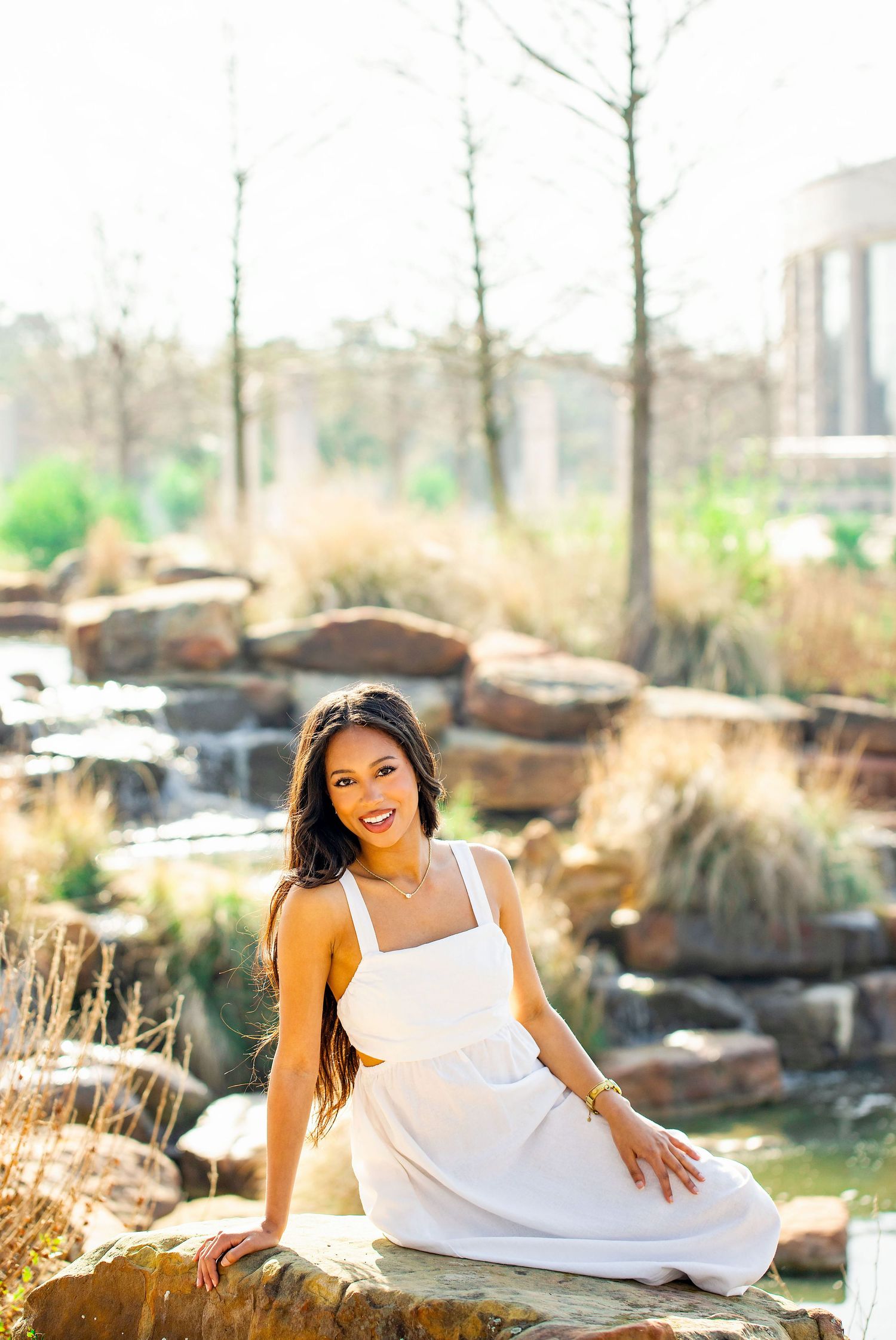 A white dress stands out against a natural backdrop of rocks and a cascading waterfall in an outdoor garden setting.