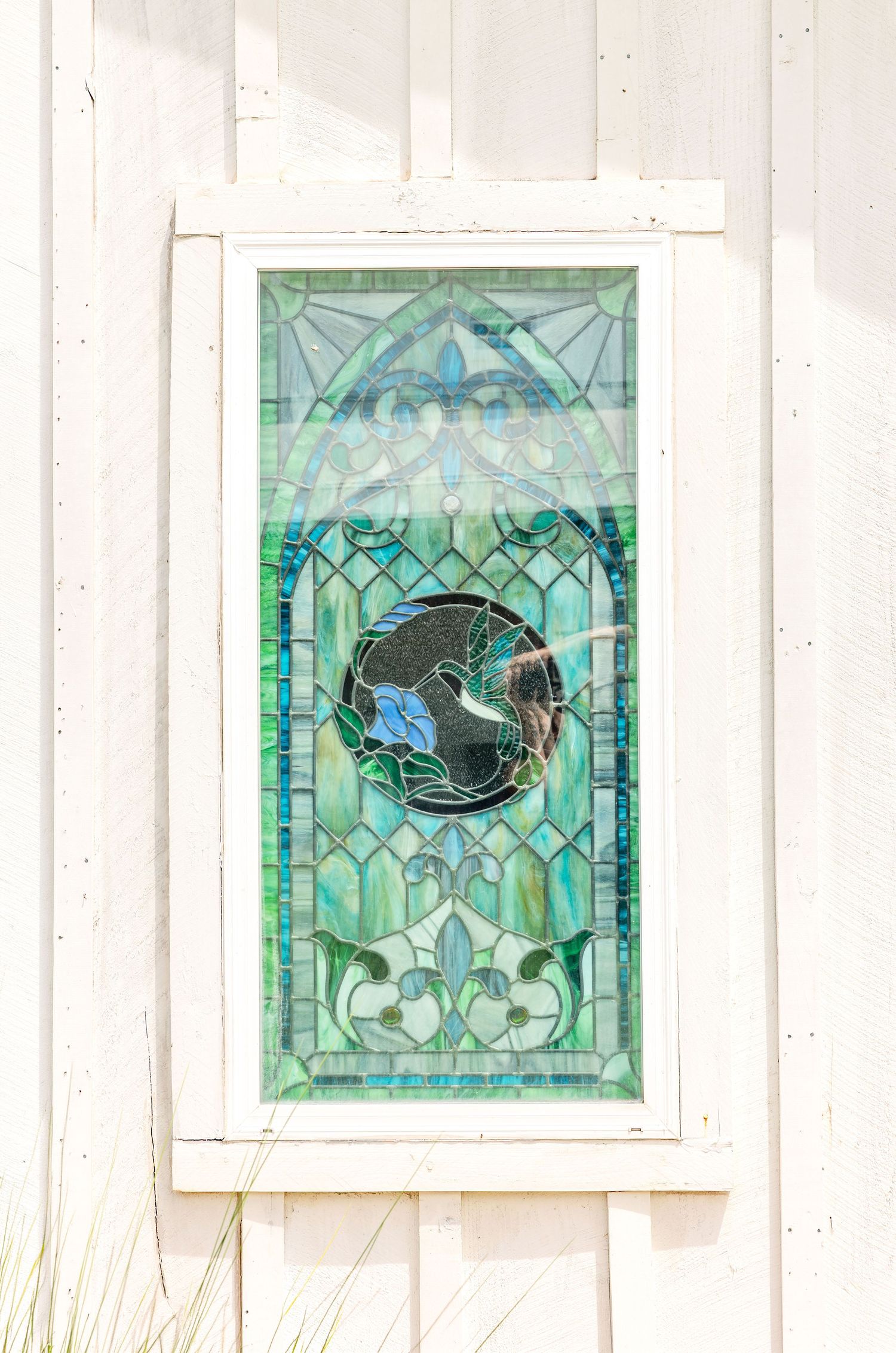 Decorative stained glass window with green and blue tones mounted on a white wooden wall exterior.