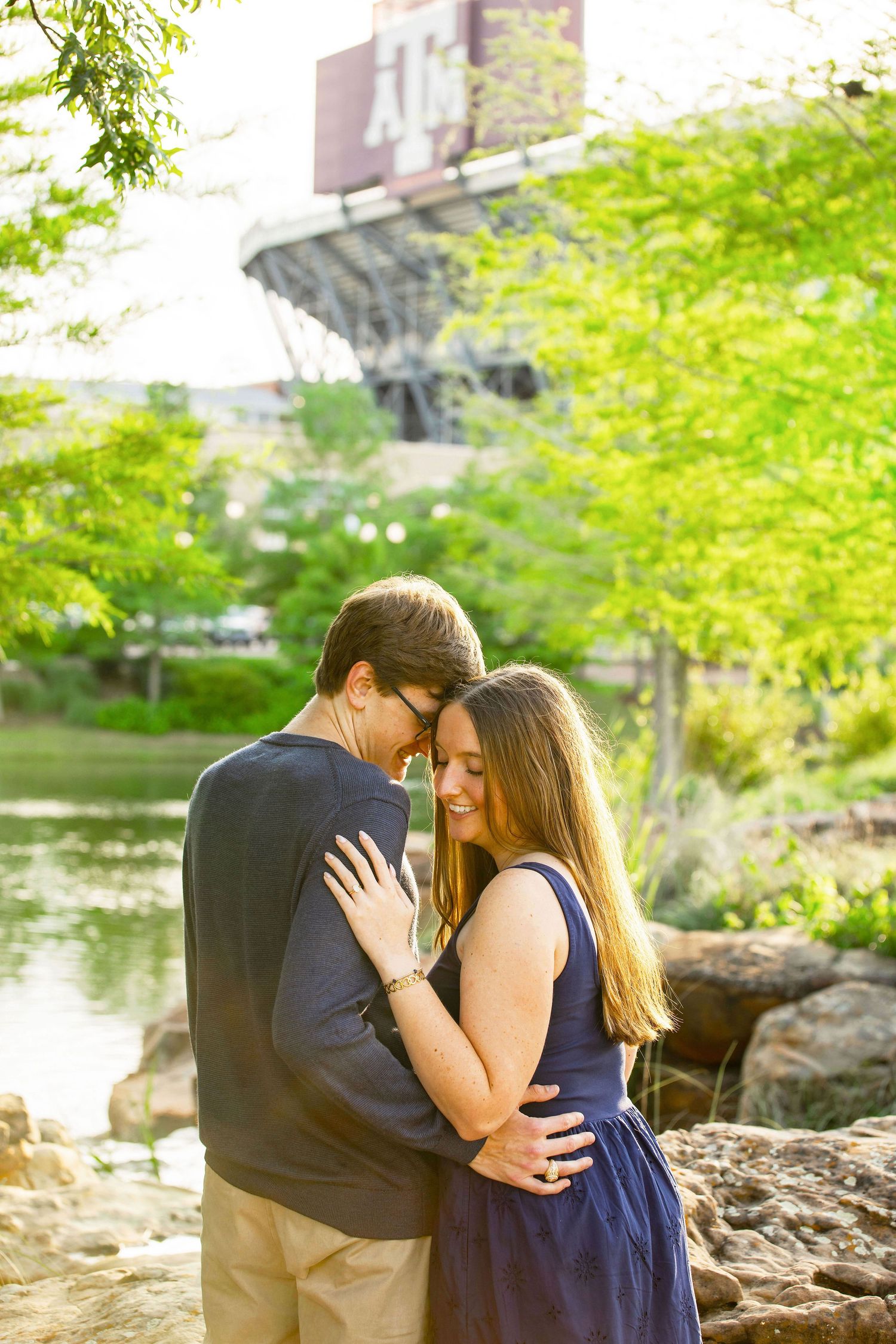 A couple embraces tenderly by a sunlit pond surrounded by lush green trees in an outdoor park setting.