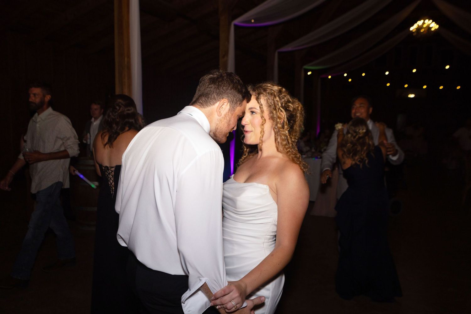 Bride and groom share an intimate slow dance at their wedding reception in a warmly lit barn venue.