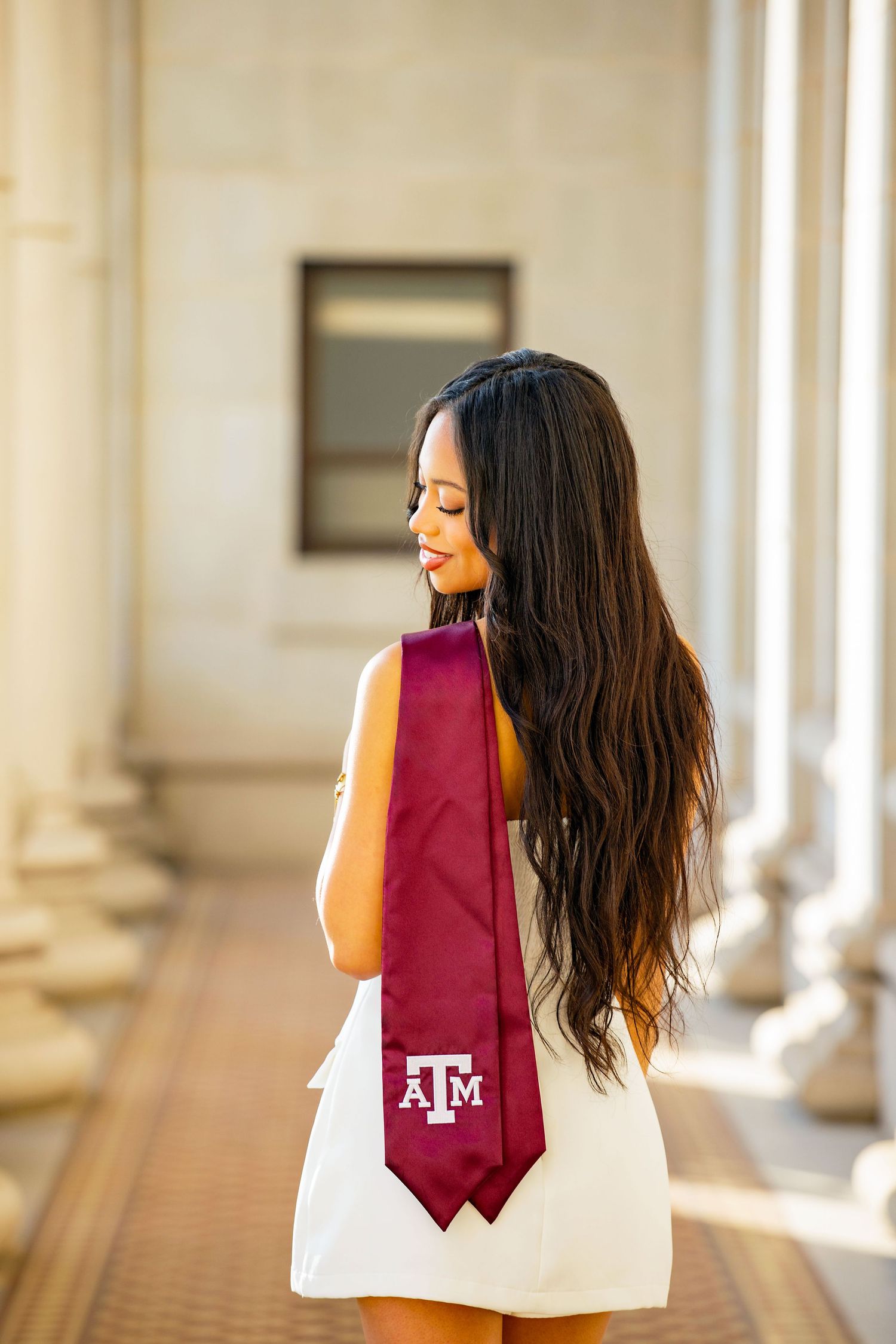 Graduate poses in white dress with maroon Texas A&M stole in front of classical columns during graduation photoshoot.