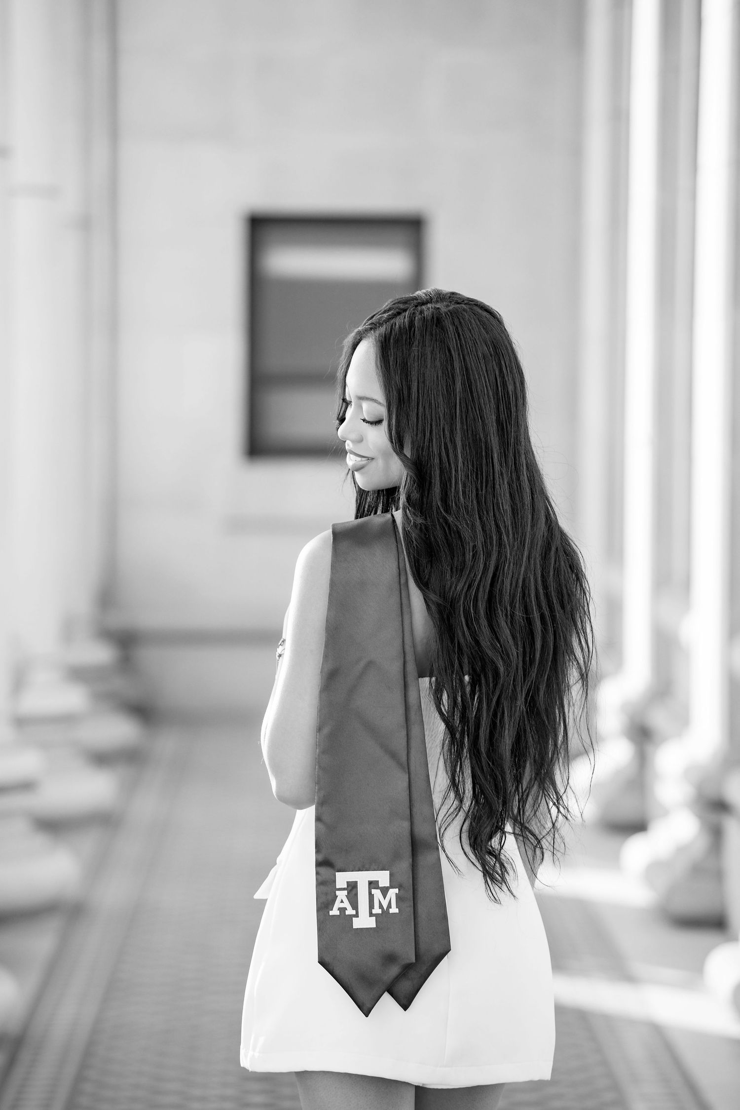 Black and white graduation photo showing long-haired graduate in white dress with Texas A&M tie viewed from behind.