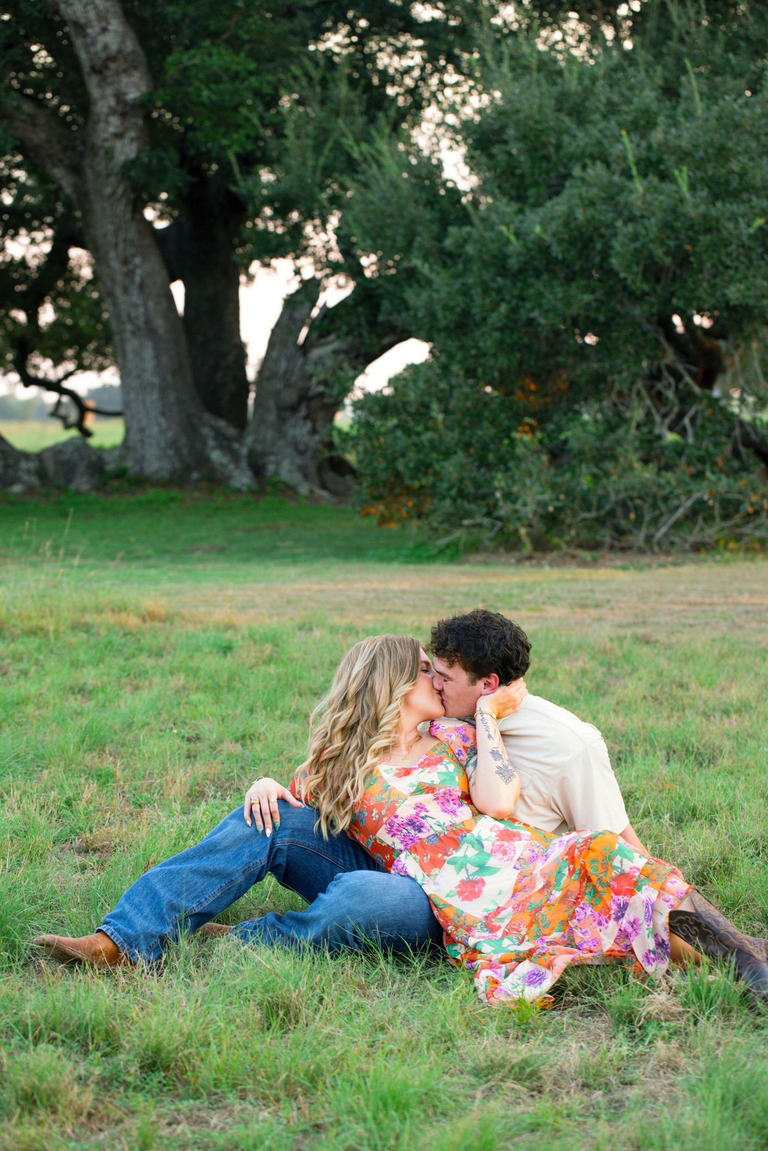 A couple lying together in a grassy field beneath large oak trees on a sunny afternoon.