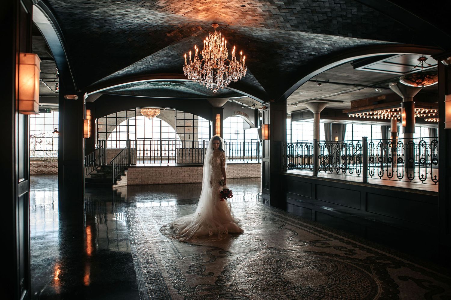 A bride in a white gown stands beneath an elegant crystal chandelier in a dark, luxurious ballroom with arched ceilings.