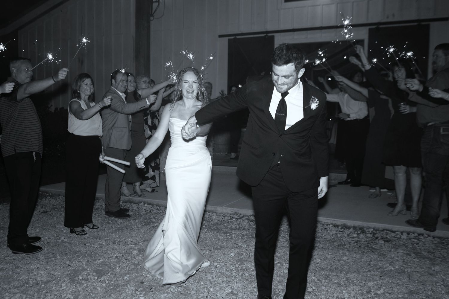 Bride and groom exit wedding reception through sparkler tunnel surrounded by cheering guests in black and white photo.