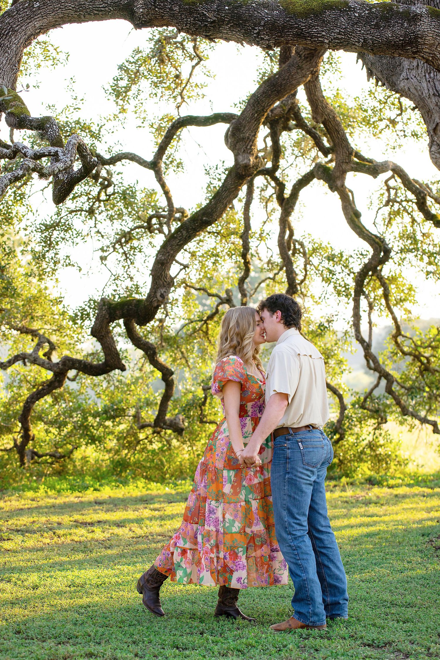 A romantic couple sharing a tender moment under a sprawling oak tree in a sunlit field.