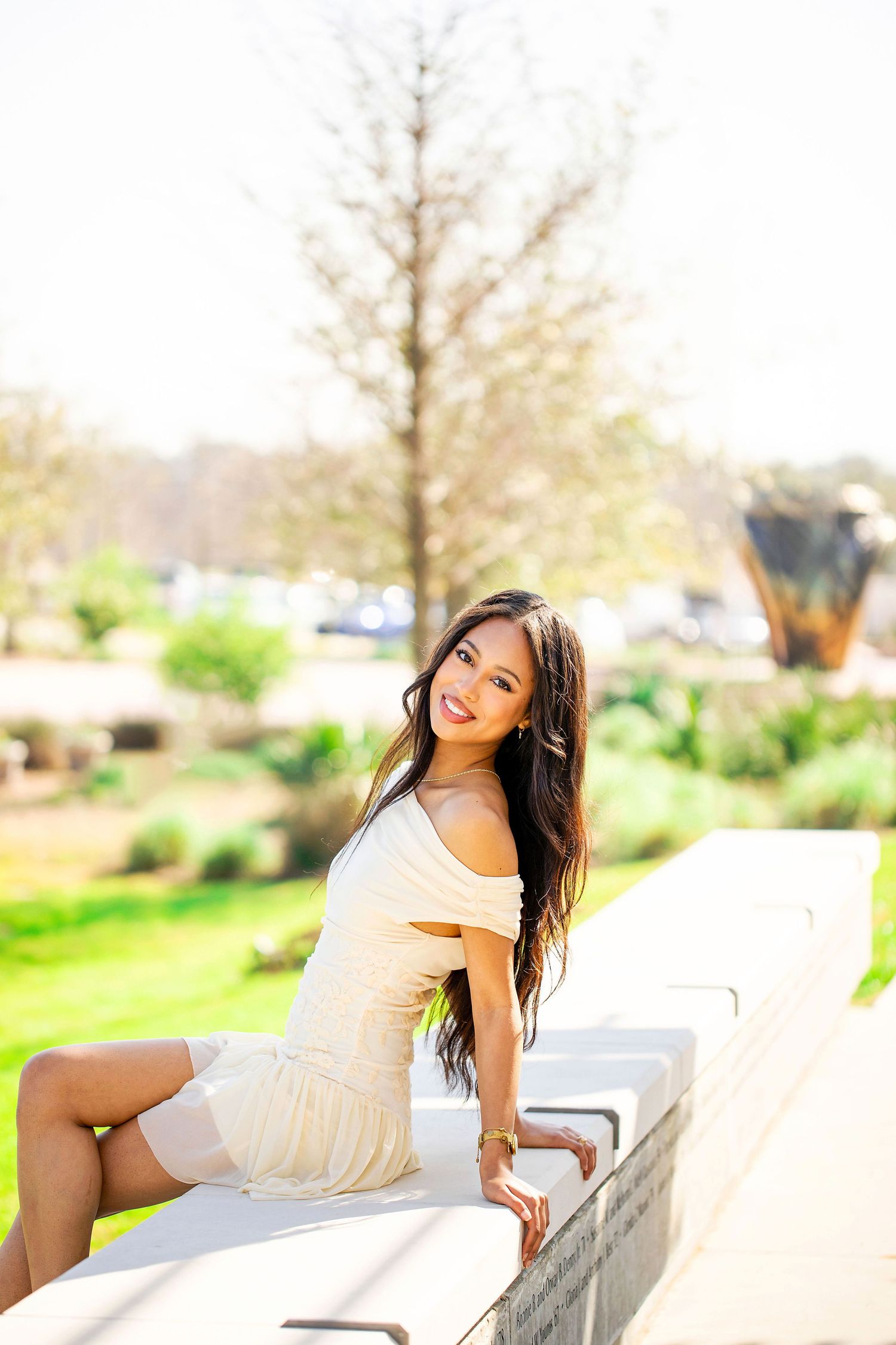 A person in a white off-shoulder dress sits casually on a concrete ledge in a sunny outdoor setting.