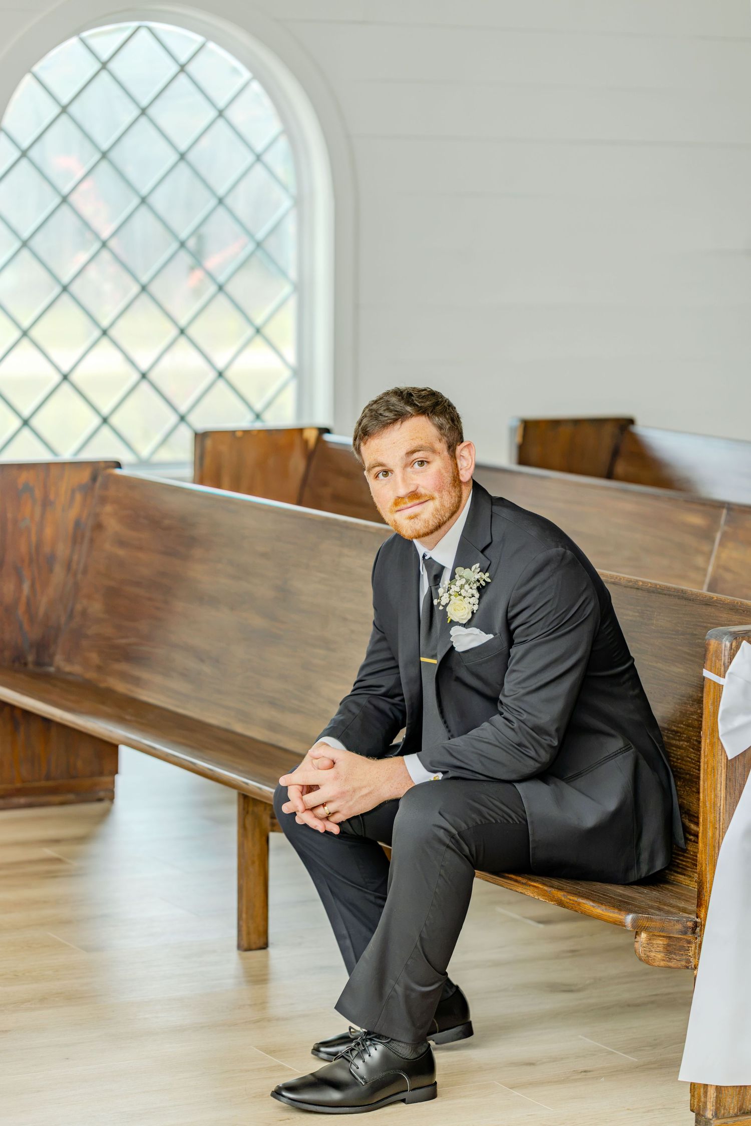 Groom in dark suit with boutonniere sits on wooden church pew near arched window before wedding ceremony.