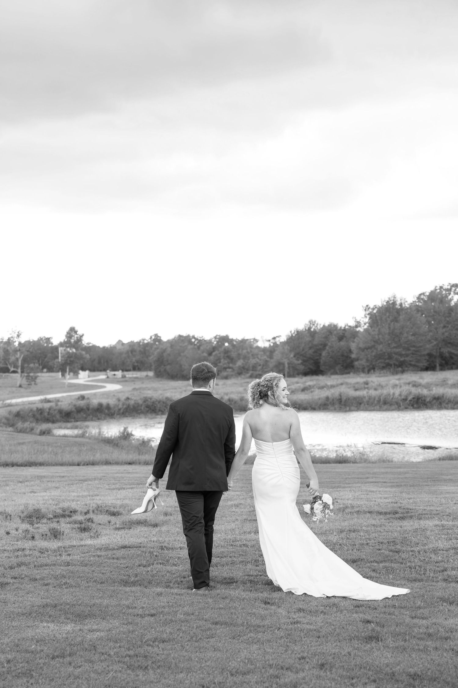 Black and white photo of a bride and groom walking hand in hand across a green field toward a pond.