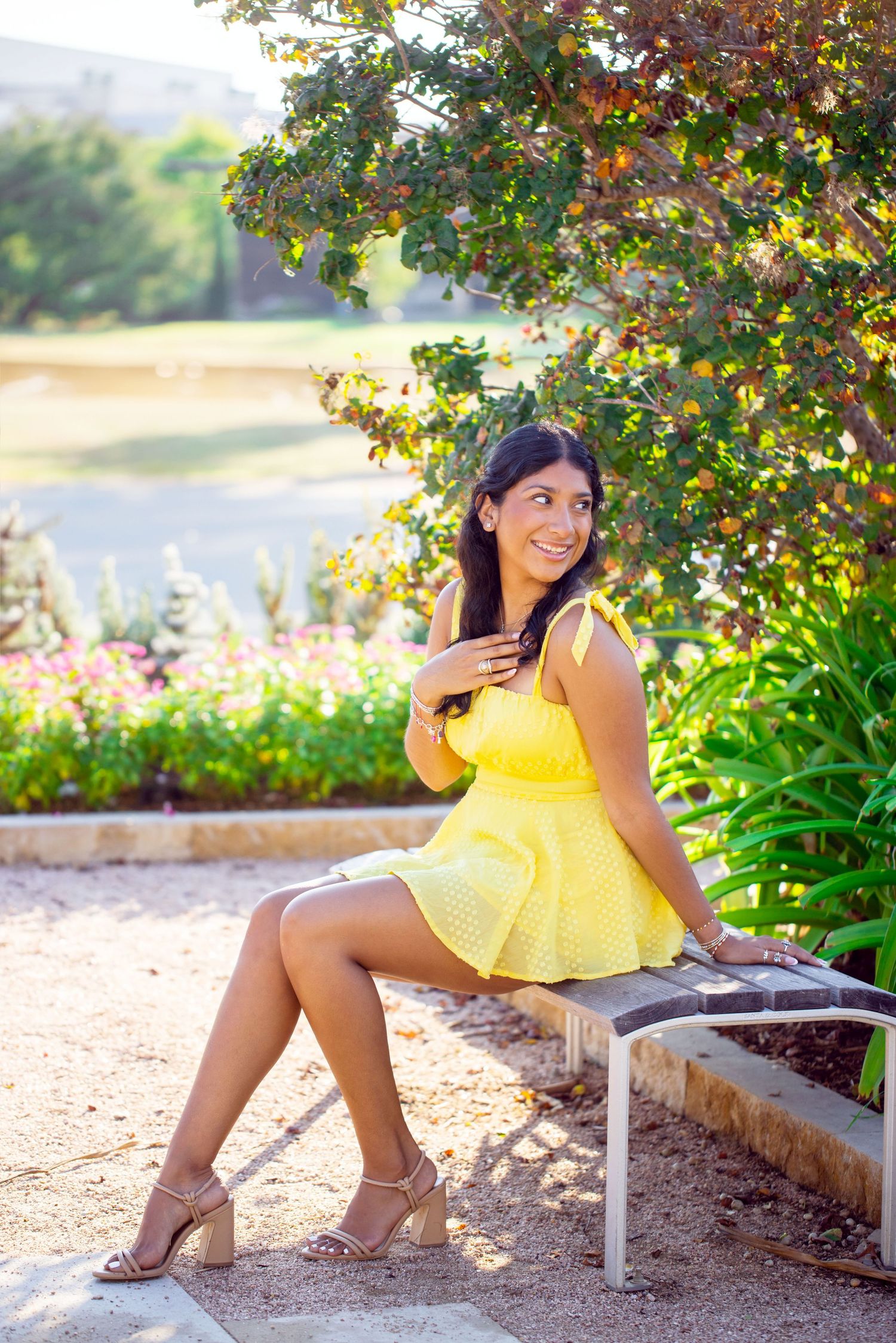 Texas A&M University graduate in pink top and white skirt captured during u in yellow sundress on garden bench with autumn foliage backdrop.