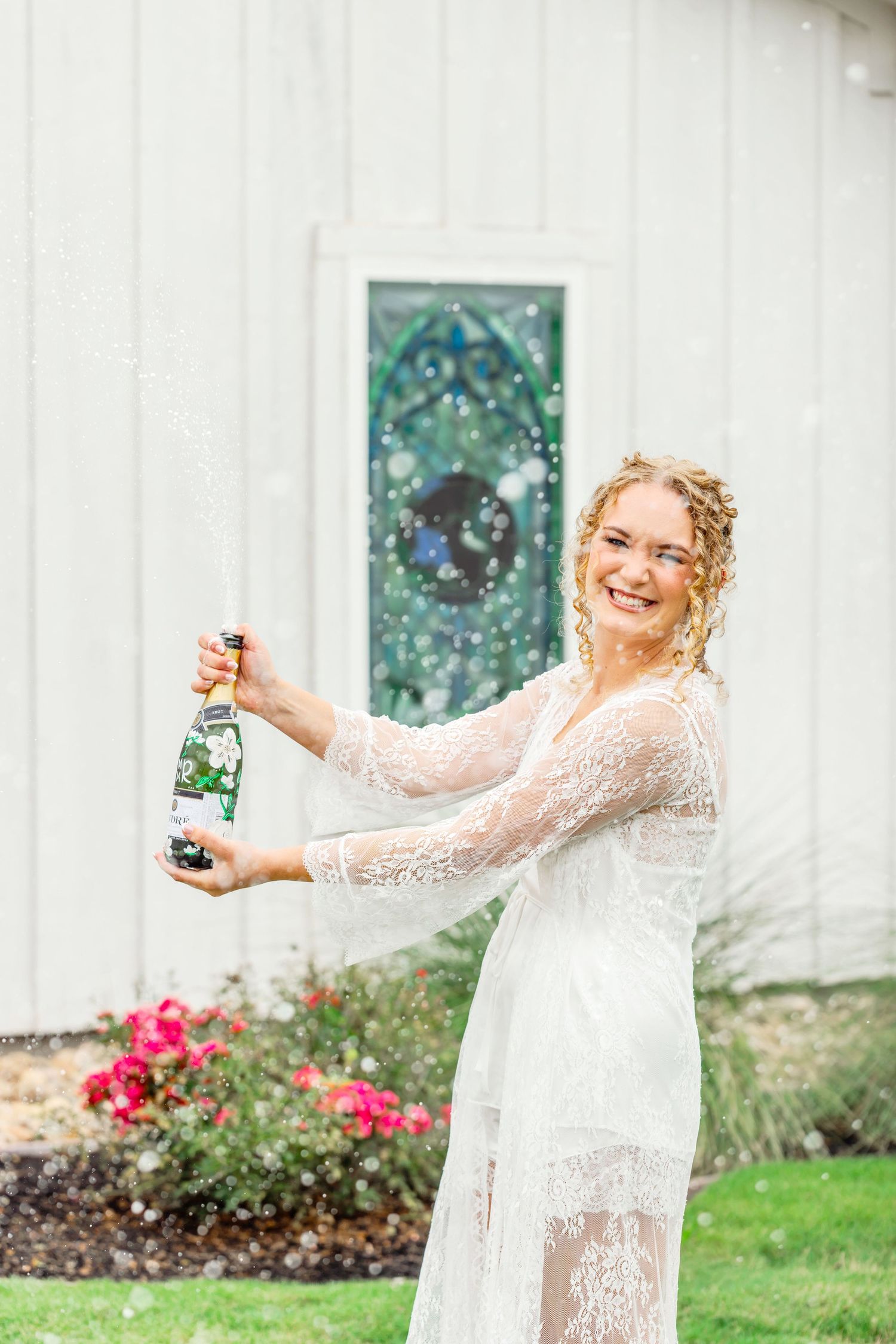 Smiling bride in white lace dress popping champagne bottle outdoors near white barn with stained glass window.