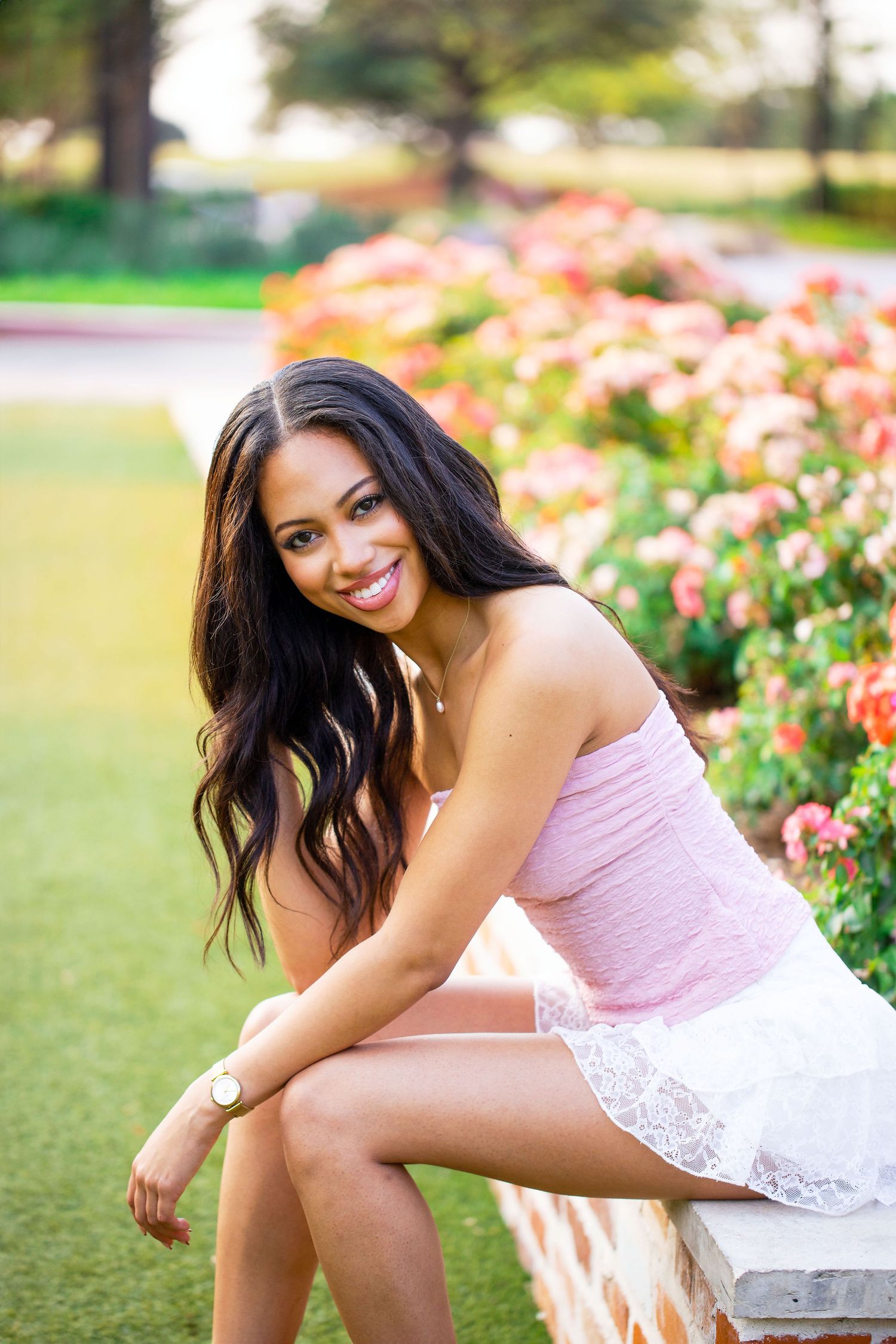 Texas A&M University graduate in pink top and white skirt photographed in Century Square in a garden setting with blooming roses.