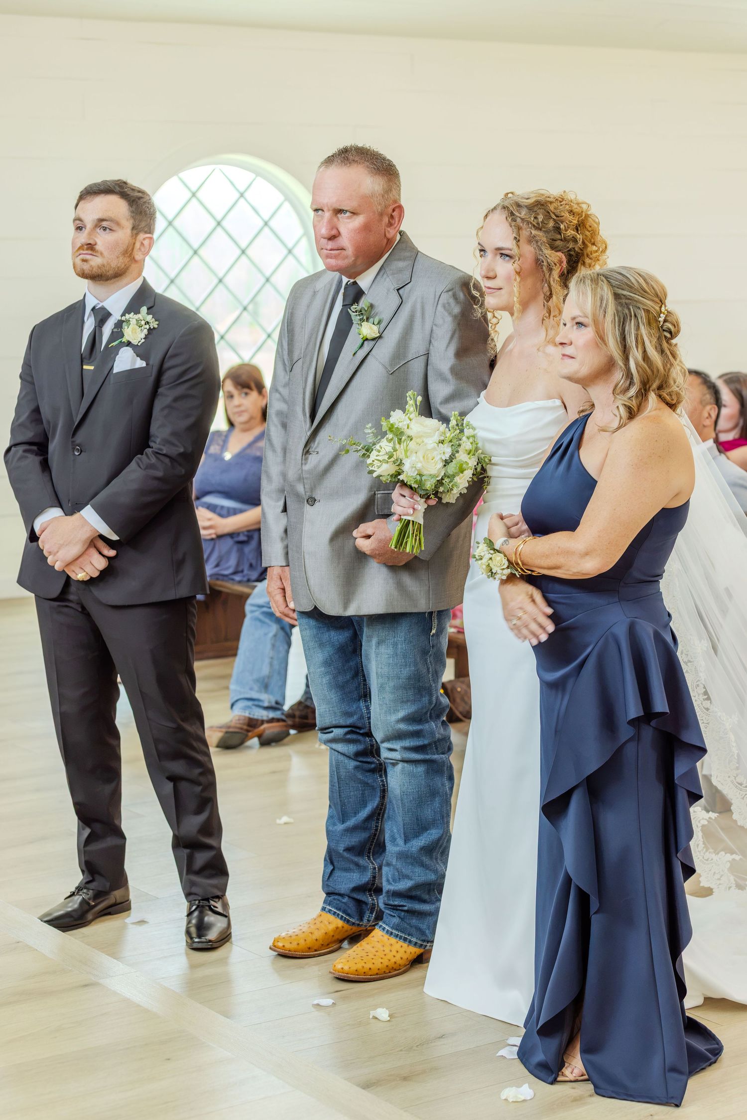 Bride in white gown holds bouquet beside father in jeans and woman in navy dress at indoor wedding ceremony.