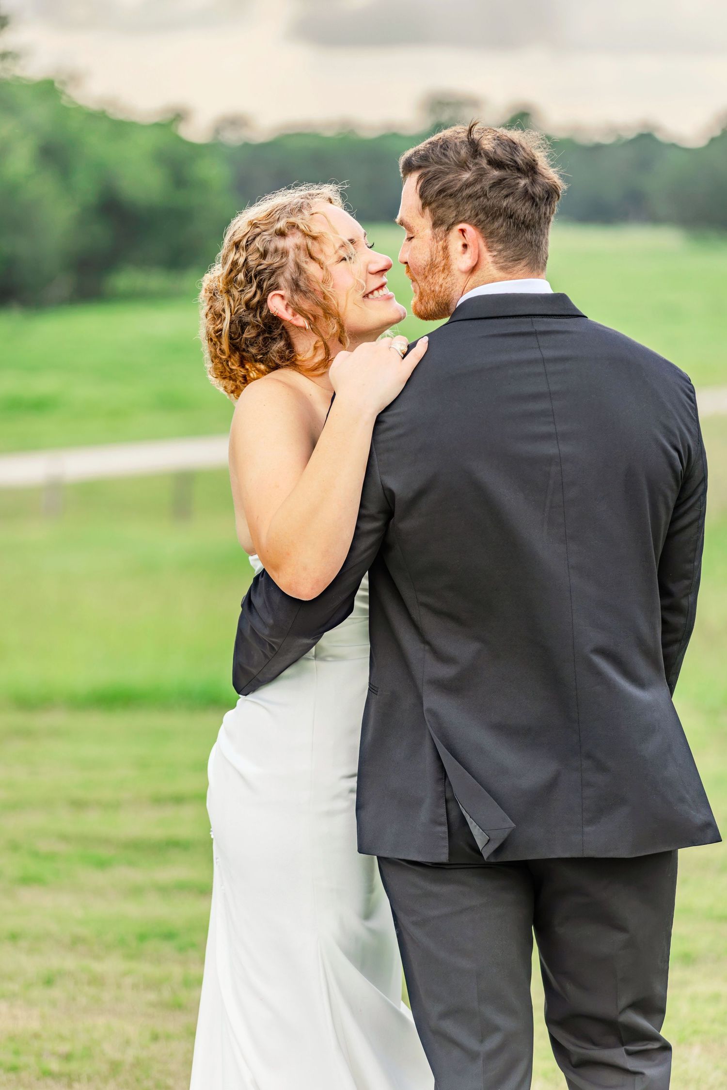 A bride and groom share a romantic moment outdoors, with the bride smiling up at her groom in a lush green field.