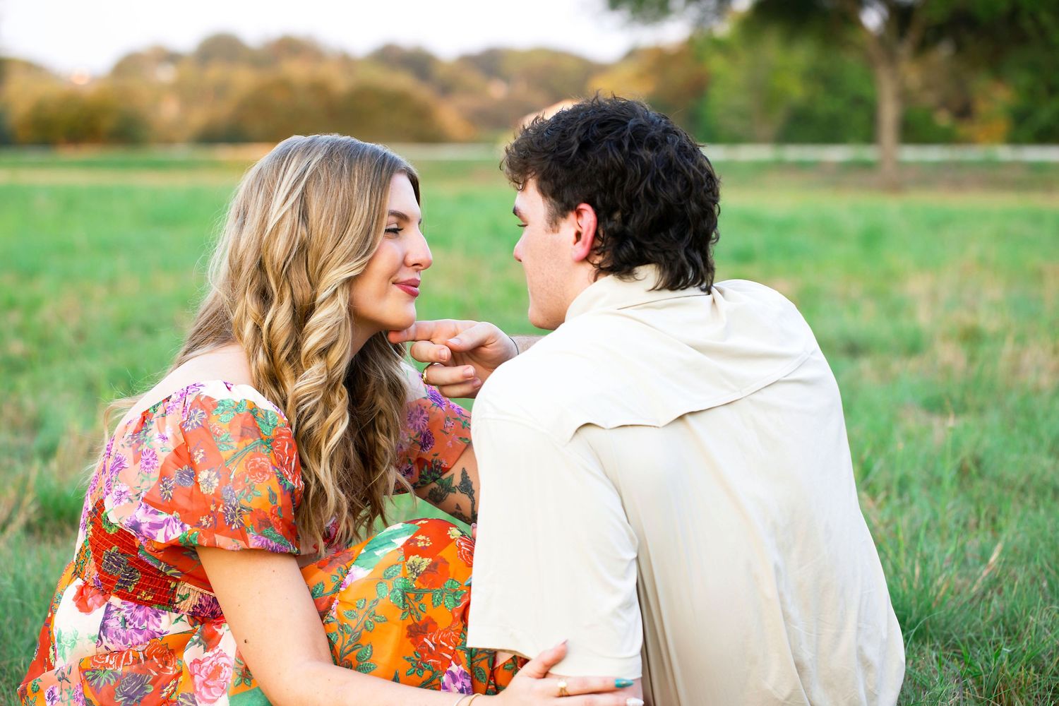 A couple sits together in a grassy meadow on an autumn day, wearing floral and white clothing.