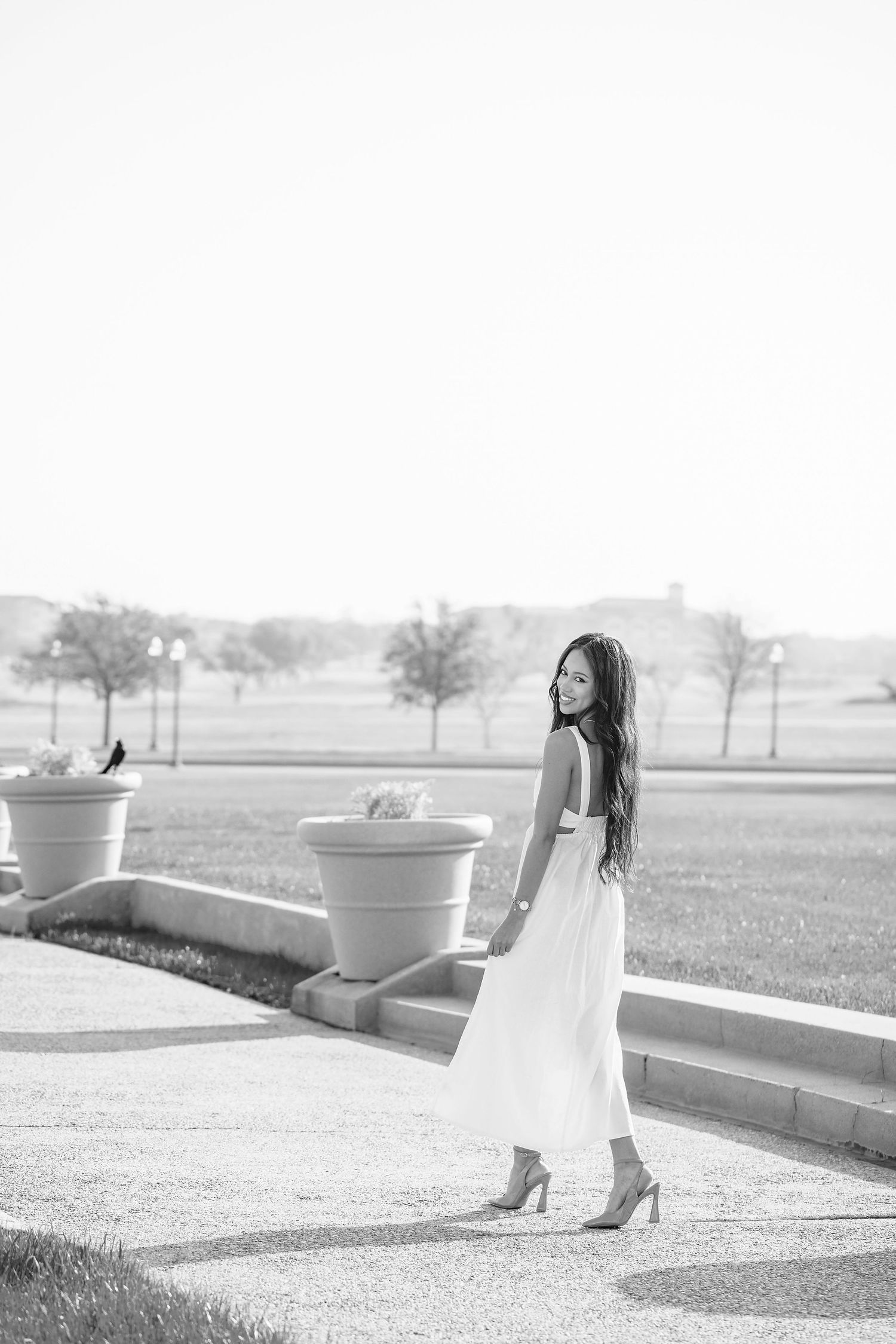 Black and white portrait of a woman in a flowing white dress standing outdoors near decorative planters.