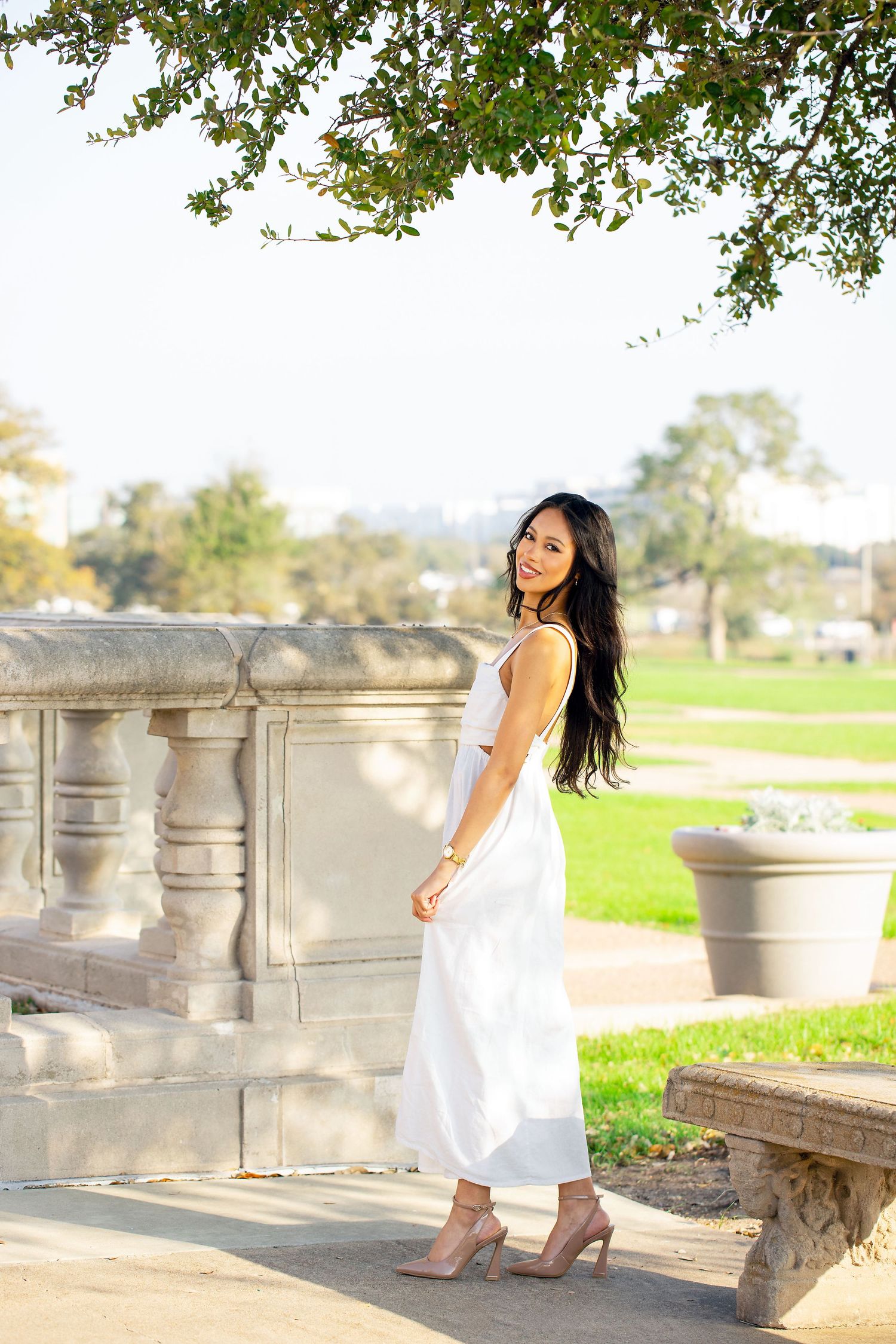 A woman in a flowing white sundress poses gracefully on stone steps in a sunny outdoor setting.