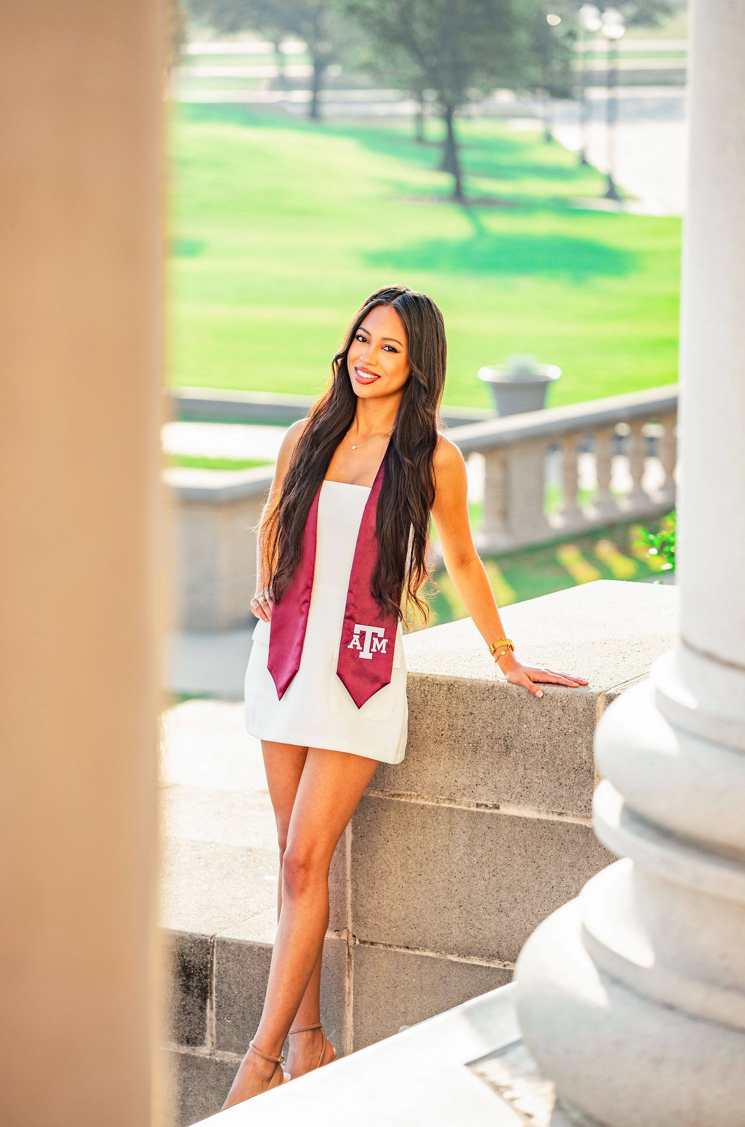 Graduate wearing white dress and maroon Texas A&M stole poses on stone steps with columns and green lawn in background.