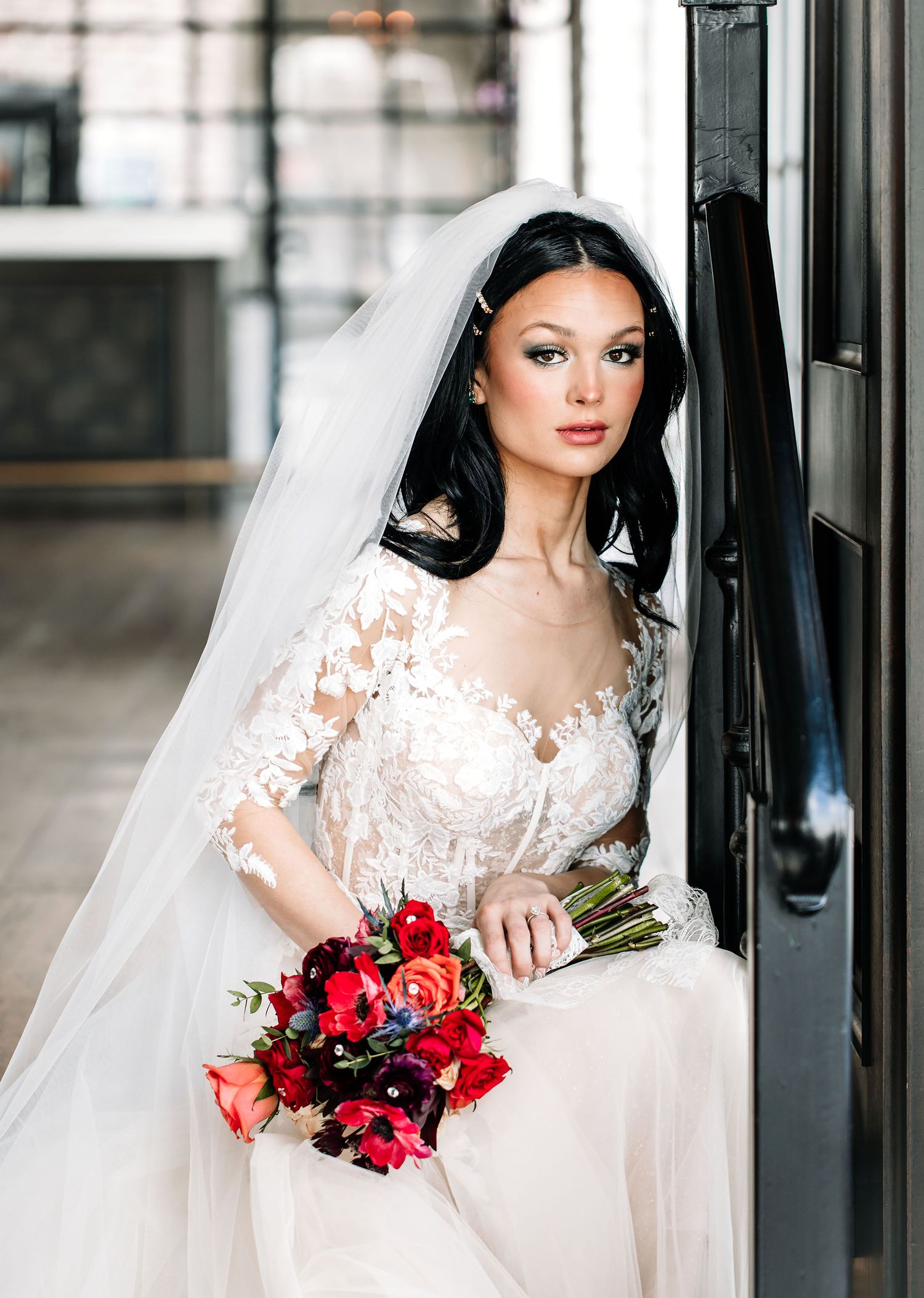 A bride in a white lace wedding dress with long sleeves holds a vibrant red and pink rose bouquet.