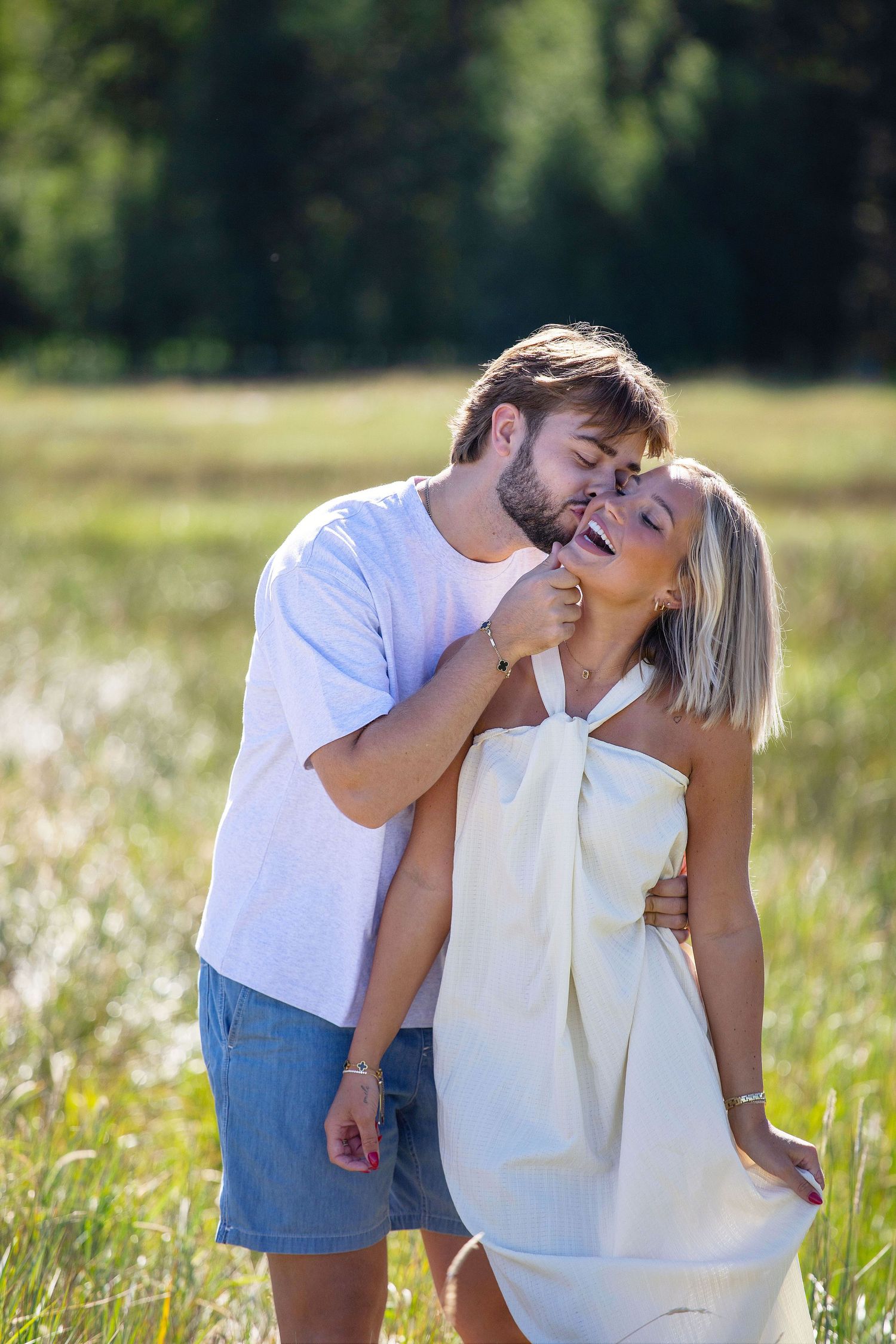 Happy couple embracing in a sunny meadow, woman in white dress laughing as man kisses her cheek.
