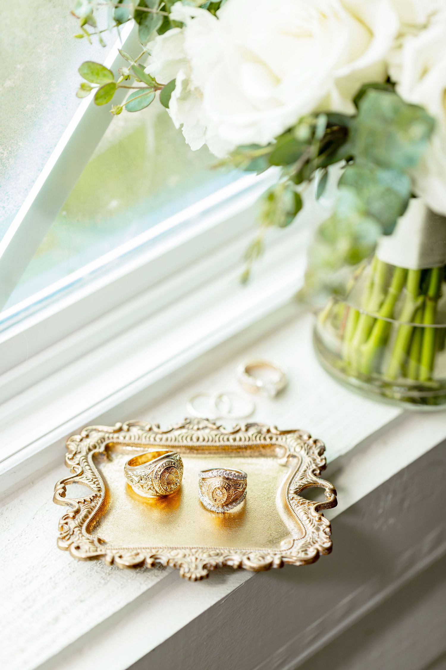 Gold signet rings on ornate silver tray by window with white roses bouquet in glass vase, elegant wedding detail.