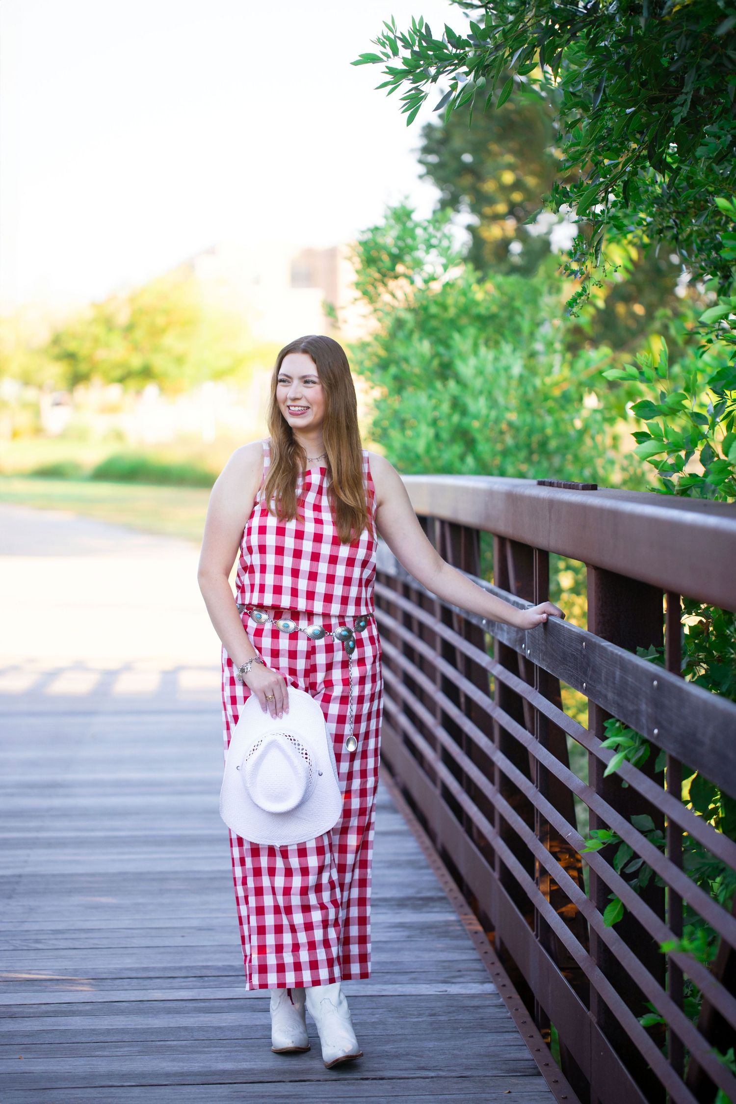Texas A&M University graduate in a red and white gingham dress photographed on a wooden bridge walkway surrounded by greenery.