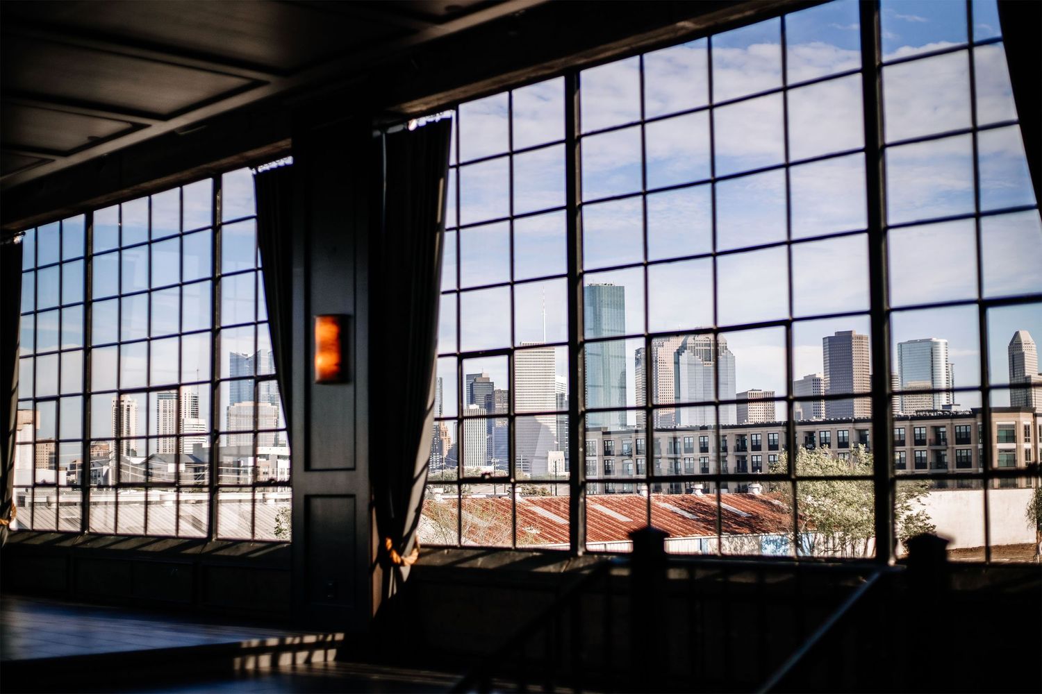 Industrial loft interior with large windows overlooking city buildings during sunset creates dramatic lighting effect.