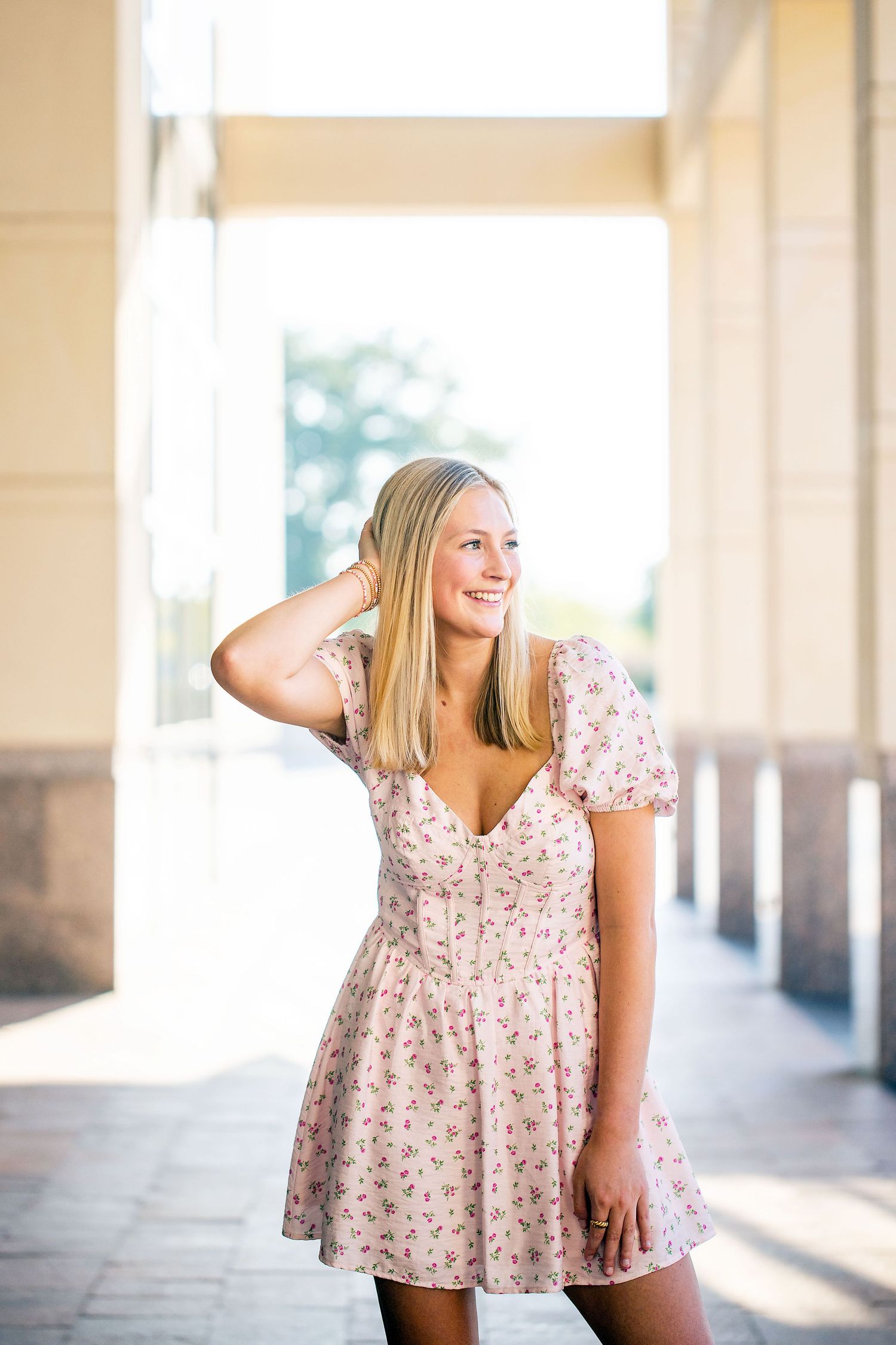 Texas A&M University graduate in pink floral dress photographed in a bright, airy corridor with natural lighting.