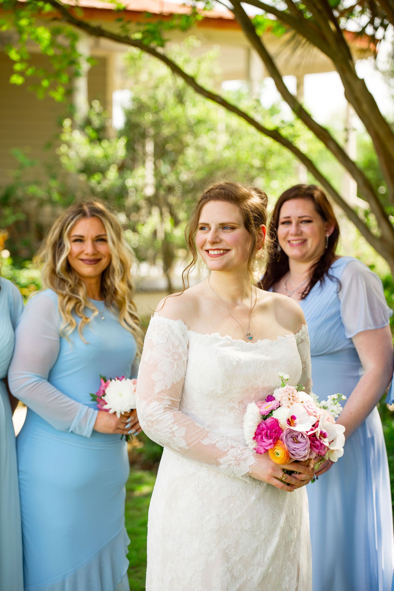 Bride in white lace gown holds colorful bouquet with smiling bridesmaids in light blue dresses outdoors.
