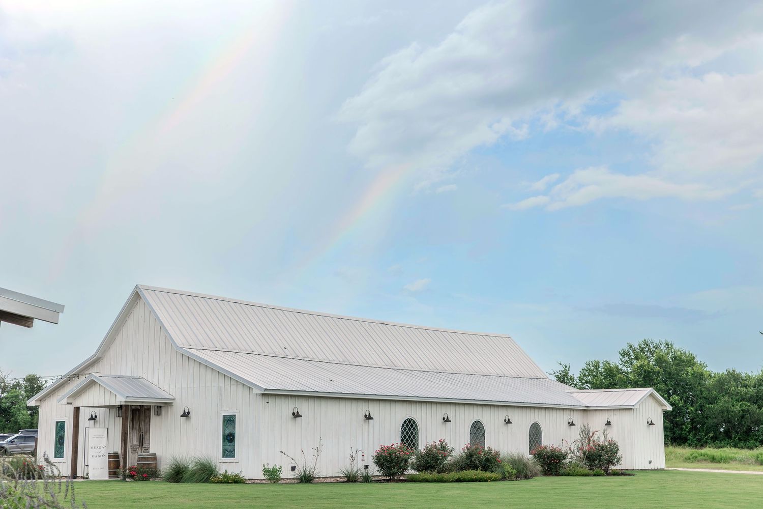 White barn venue with metal roof under a rainbow and blue sky, surrounded by green trees and manicured lawn.