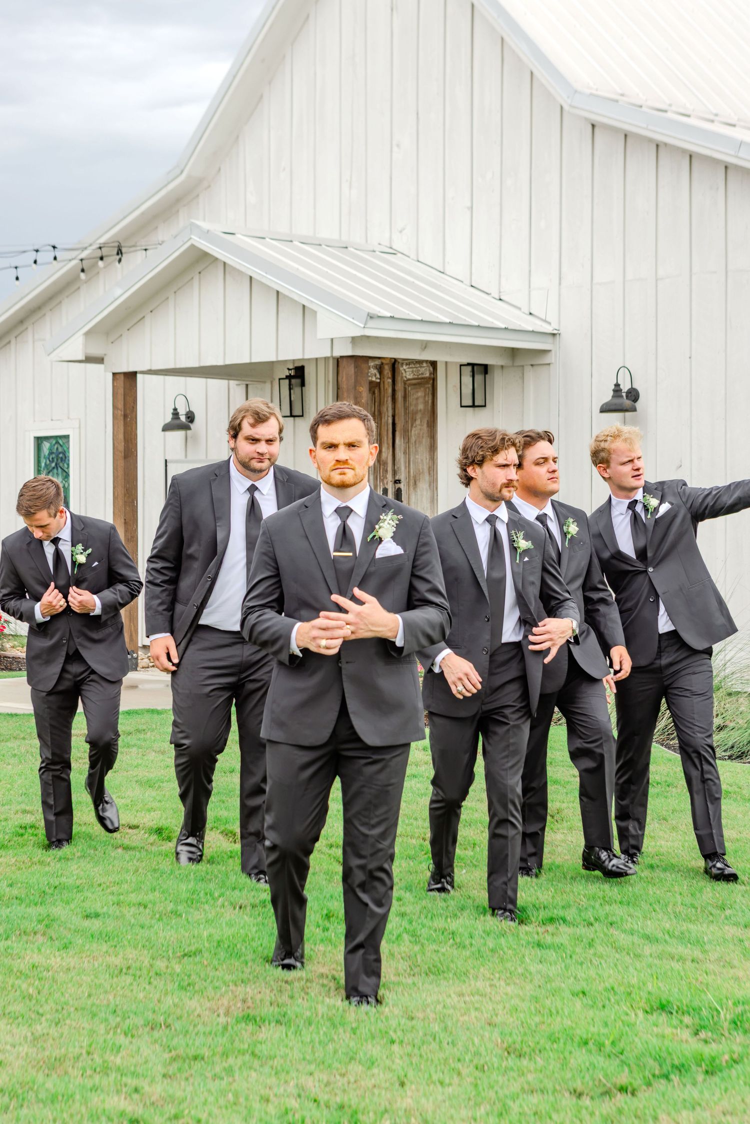 Groomsmen in dark suits with boutonnieres walk confidently across green lawn in front of white barn wedding venue.