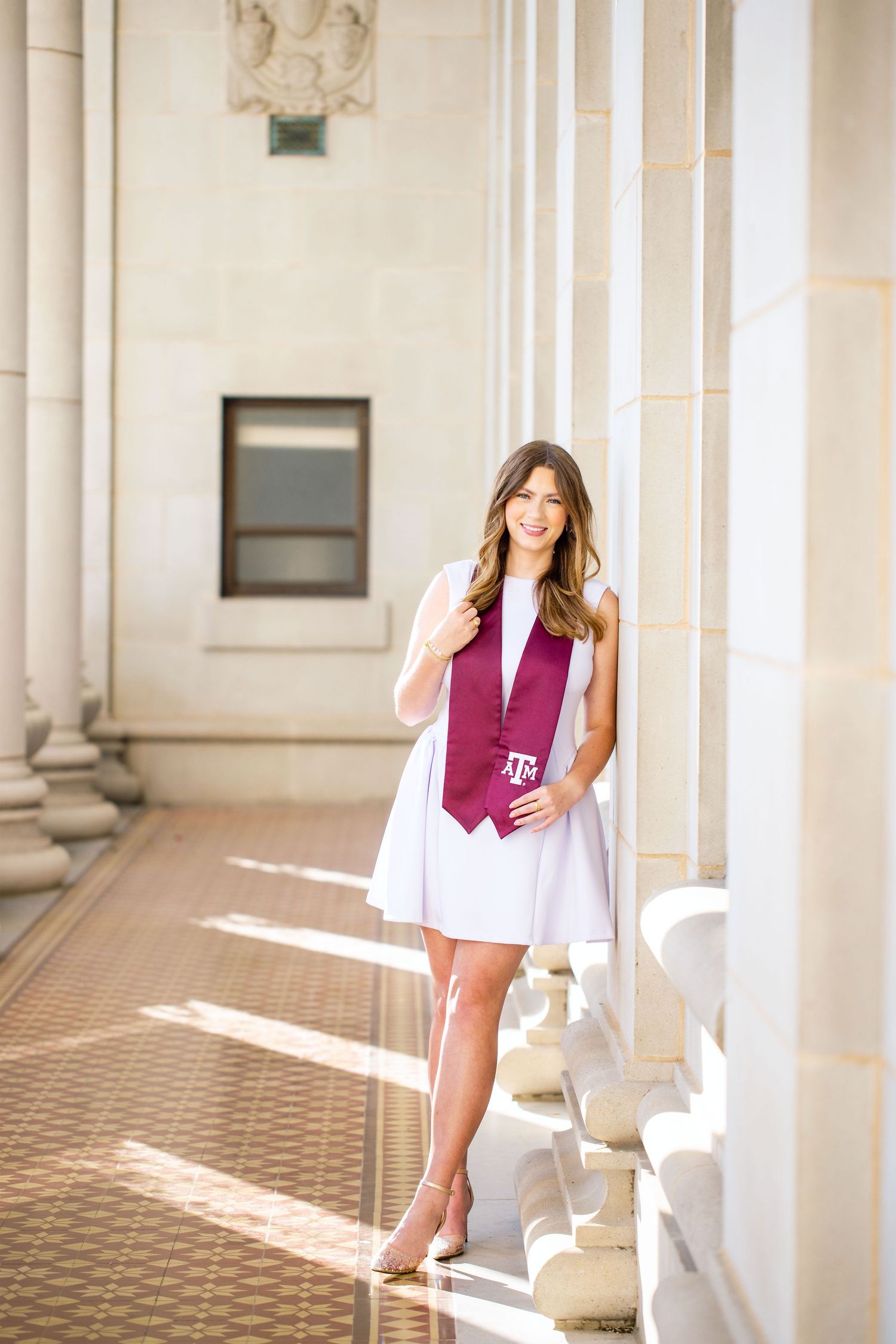 A young woman with long brown hair wearing a pink top and white skirt stands in front of a building with columns.