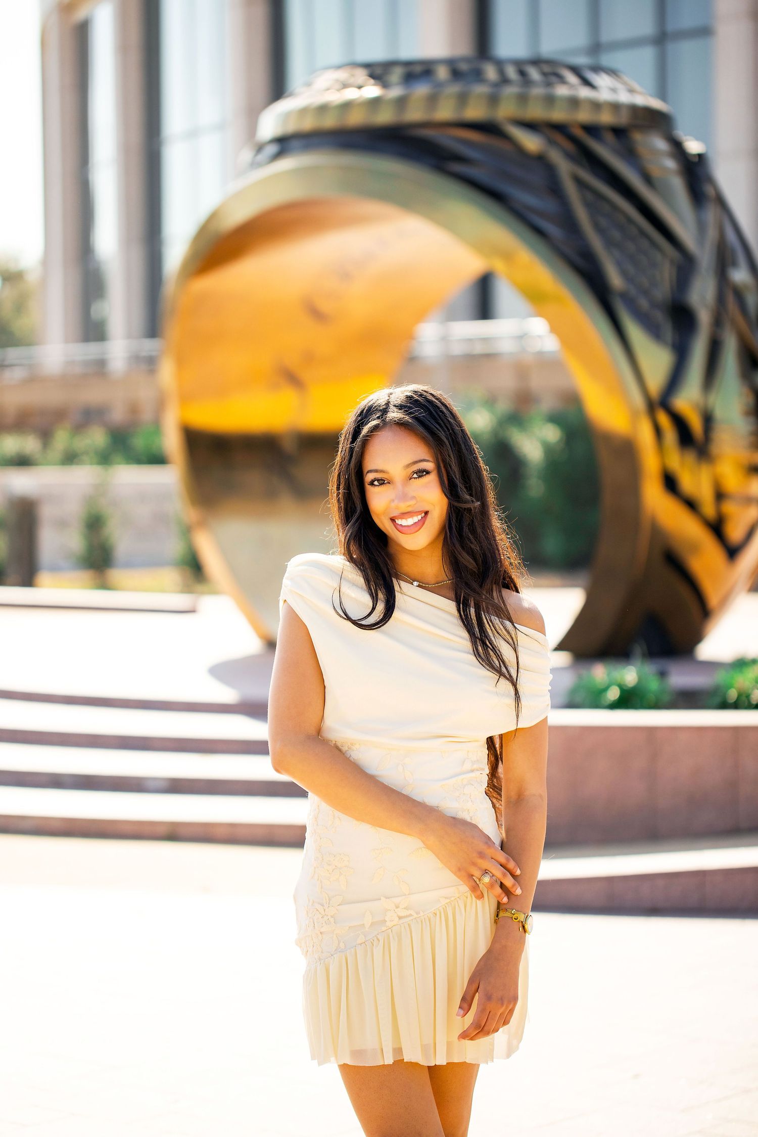 Woman in white ruffle dress poses outdoors in urban setting with golden hour lighting and architectural elements.