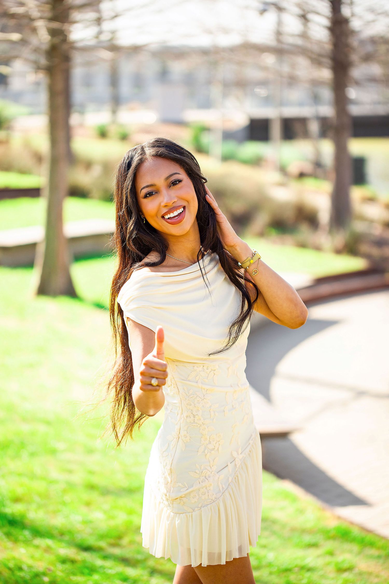 A person in a white dress poses joyfully outdoors on a sunny day with trees and grass in the background.