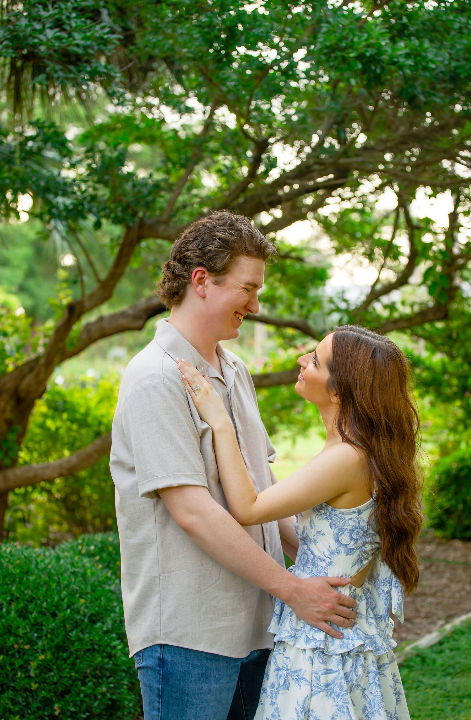 A smiling couple embraces outdoors in a lush green garden, gazing at each other during an engagement photo session.