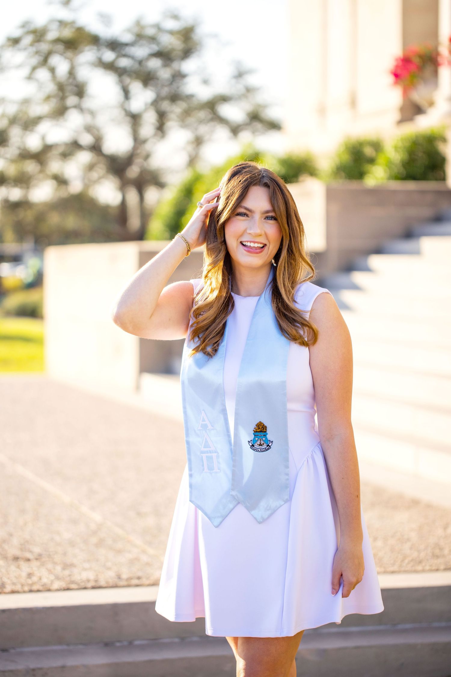 A smiling young woman with long brown hair stands outdoors on a sunny day.