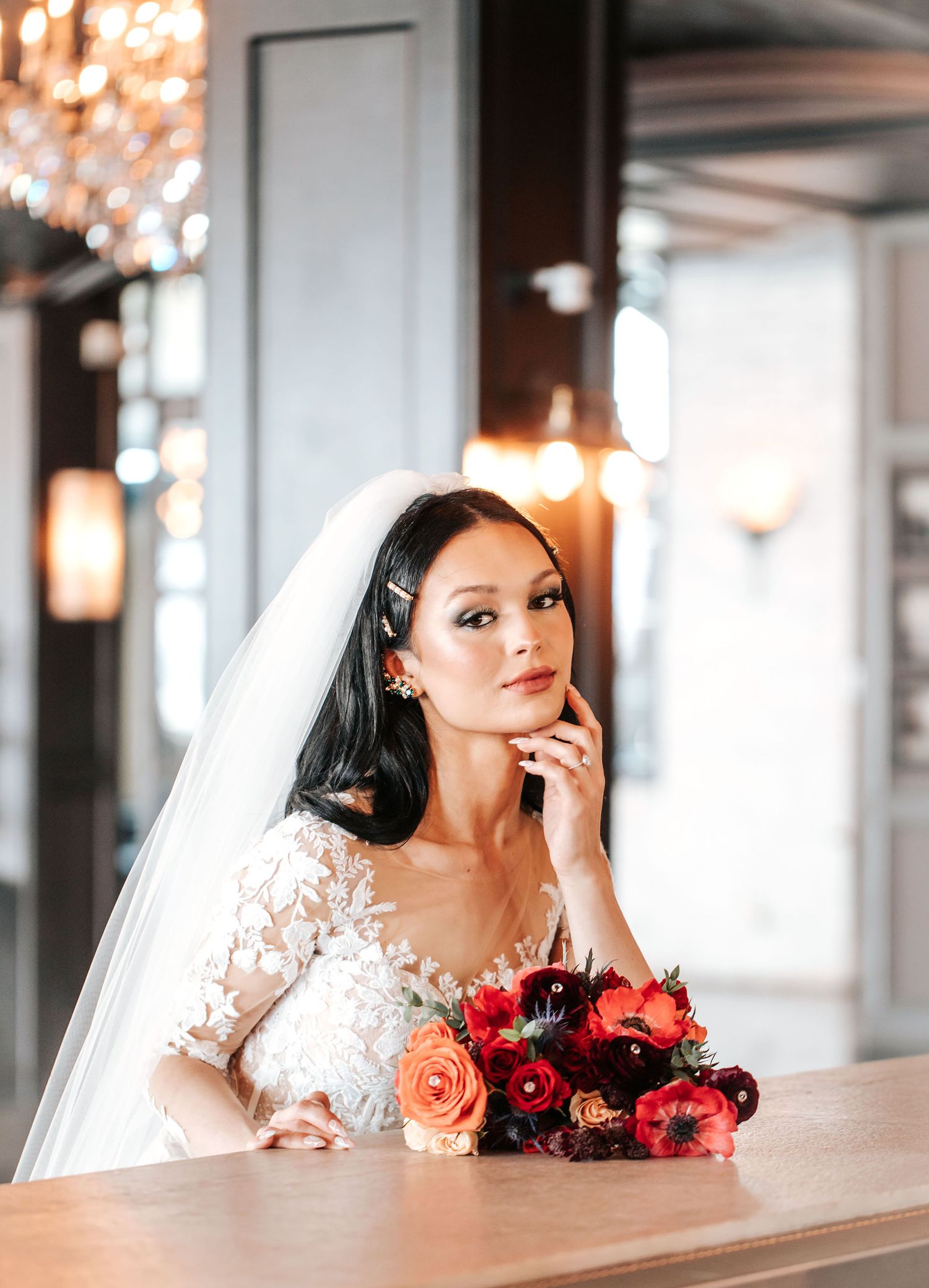 A bride in a white wedding dress poses with a vibrant red and coral bouquet against an elegant indoor backdrop.