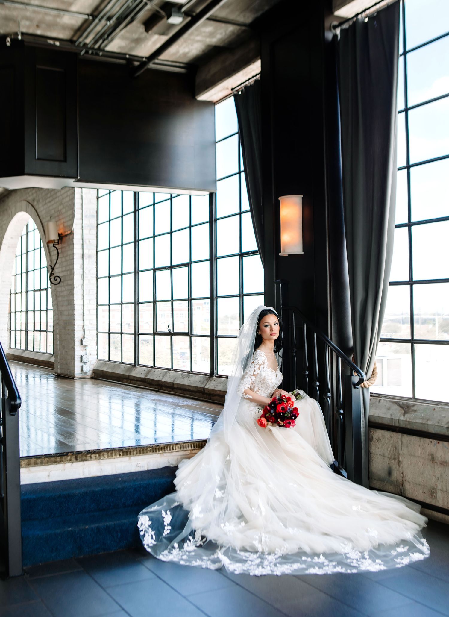 A bride in a flowing white lace gown poses gracefully in an industrial loft space with large windows and natural light.